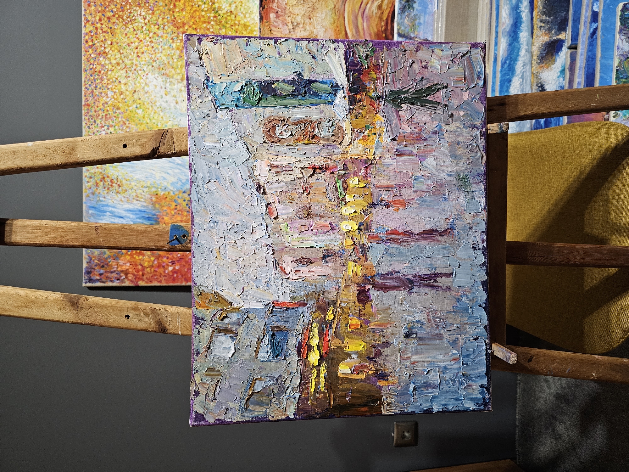 An eye-level indoor shot depicts an impressionistic painting of a street in Plovdiv, Bulgaria, rendered with thick, textured brushstrokes in a palette of blues, grays, purples, yellows, and oranges. The painting is displayed on a wooden rack system with horizontal slats, positioned in front of a gray wall and other artworks, one featuring abstract orange and yellow hues and another with blue vertical lines. A yellow chair is partly visible to the right of the painting. A dark gray wall plug is partially visible beneath the lower edge of the painting.