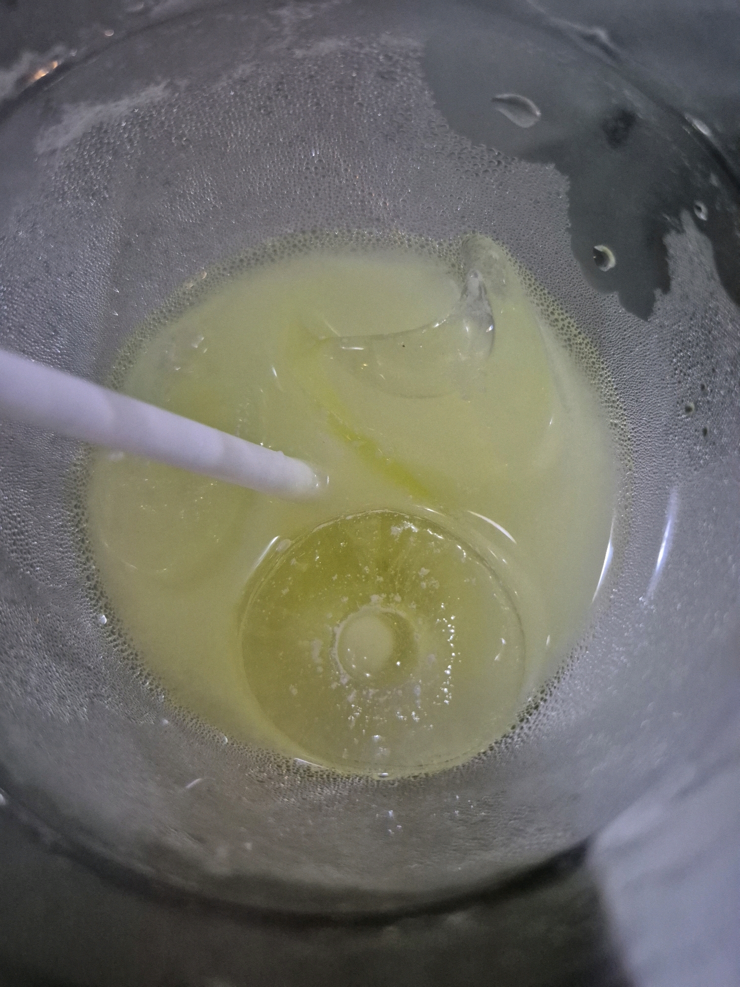 A top-down, close-up shot shows a clear glass filled with a pale yellow liquid, likely a beverage, and several ice cubes. A white straw enters the glass from the left side, submerged in the liquid. The exterior of the glass is covered in condensation, indicating it is cold. The lighting is somewhat dim, with reflections visible on the glass and ice. The scene appears to be indoors, with a dark, out-of-focus background. There are no people or animals visible in the image. The location in Pabelan, Indonesia, is not discernible from the visual information provided.