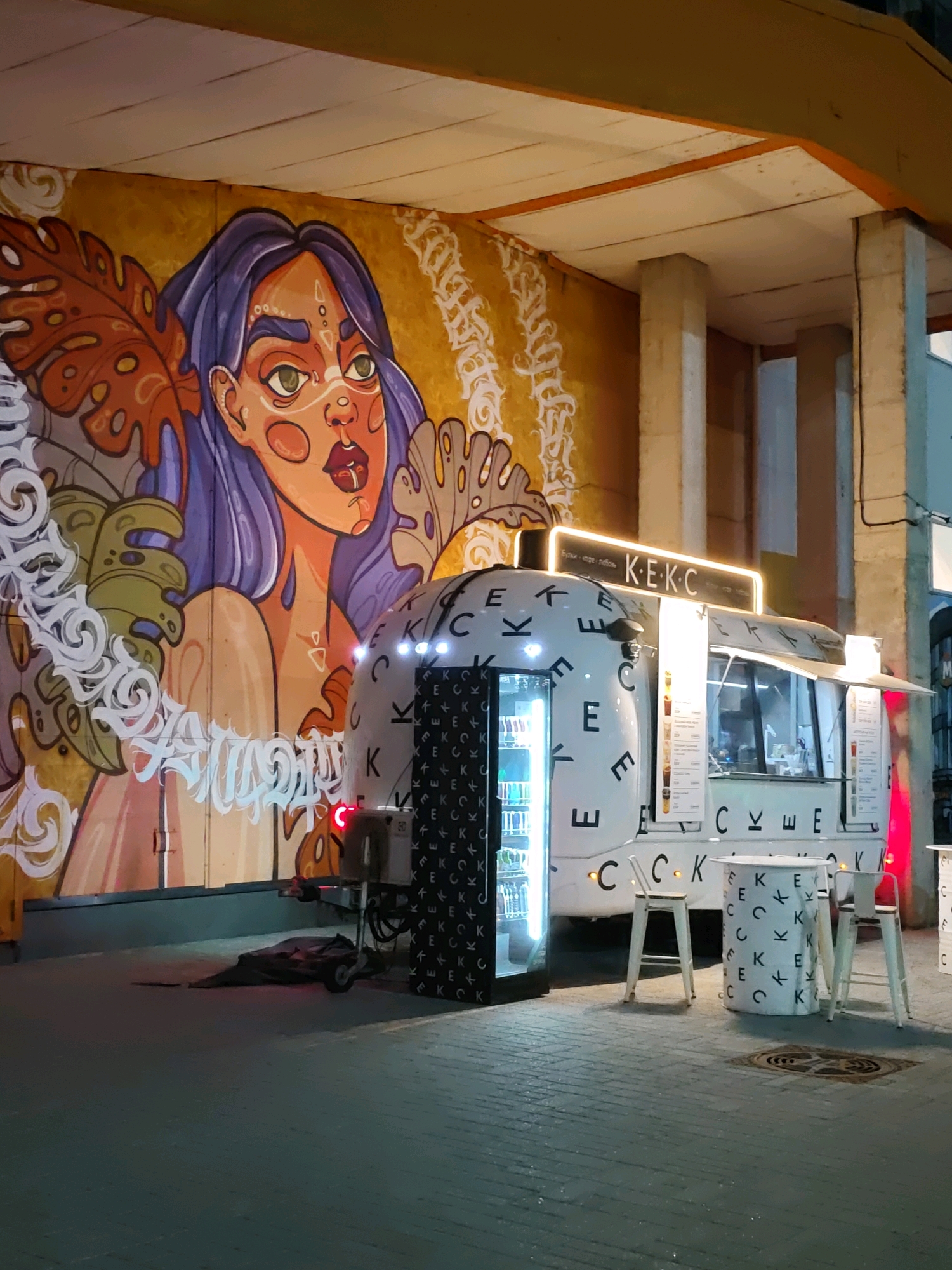 An outdoor food truck, branded with "K.E.K.C.", is parked in front of a large mural of a stylized woman with purple hair. The food truck has a bright, illuminated sign with Cyrillic text above its service window. White tables and stools, also decorated with the "K.E.K.C." logo, are set up in front of the truck. The scene appears to be a street or public area in Rostov-on-Don, Russia, possibly a pedestrian zone or square, as indicated by the pavement and surrounding infrastructure. The time appears to be evening or night, as artificial lighting is prominent. There are no people visible in the frame.