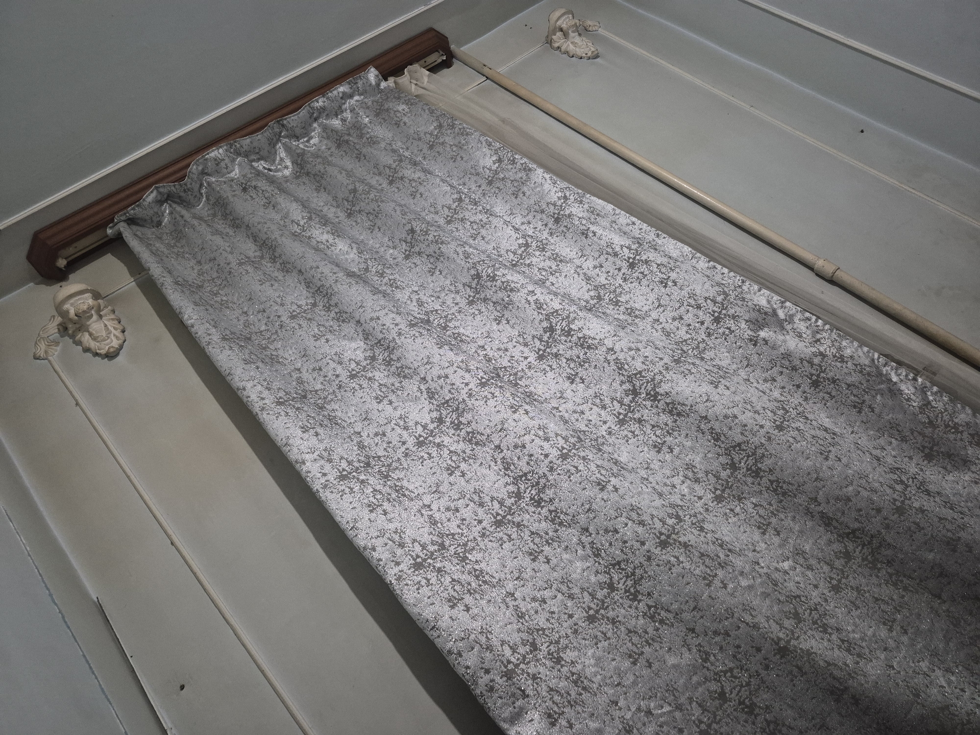 The image displays an indoor scene, primarily focusing on a closed curtain and a section of a wall. The perspective is an overhead, angled shot looking down.
The main subject is a silver-grey curtain, appearing to be made of a crushed velvet or heavily textured fabric with a mottled, abstract pattern. It hangs from a dark brown, possibly wooden, curtain pelmet or valance that conceals the curtain track. The fabric has visible folds and gathers along the top.
Flanking the curtain pelmet on the light blue-grey wall are two ornate, off-white decorative elements, possibly plaster or carved wood corbels. The wall itself features horizontal lines, suggesting either paneling or architectural molding.
To the right of the curtain, resting against the wall, is a long, slender, light beige pole, possibly a curtain draw wand or a decorative rod. The floor or lower wall area visible beyond the curtain and the pole is also in a similar light blue-grey tone, with subtle tonal variations and lines consistent with interior wall surfaces.
There are no people or discernible activities taking place. No visible text is present in the image. The lighting suggests an indoor setting, but the time of day and specific location cues (beyond an interior room) are not explicitly visible.