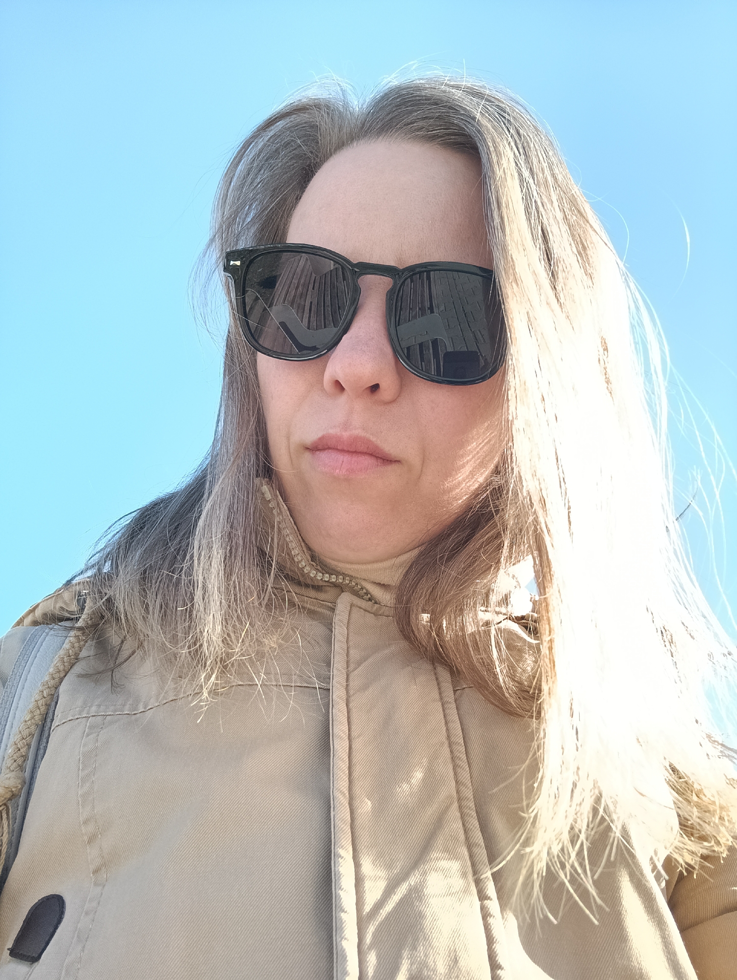 A close-up, low-angle portrait captures a woman outdoors against a bright, clear blue sky, likely in Chaikovsky, Russia. She wears dark, square-framed sunglasses that cover her eyes, with a noticeable crack on the left lens. Reflections in the sunglasses reveal vertical patterns, possibly from nearby buildings or structures. The woman's expression appears neutral to slightly pensive, with her lips slightly parted. Her hair, a blend of blonde and grey, frames her face; the hair on her right side is intensely lit by direct sunlight, creating a bright, almost glowing effect. She is dressed in a beige or khaki-colored zippered jacket, beneath which a lighter-colored turtleneck collar is visible. Details of the jacket include a braided drawstring on the left shoulder and a small, dark, possibly leather patch on the lower left. The bright, cloudless sky and strong sunlight suggest it is daytime, under clear weather conditions. No visible text is present in the image.