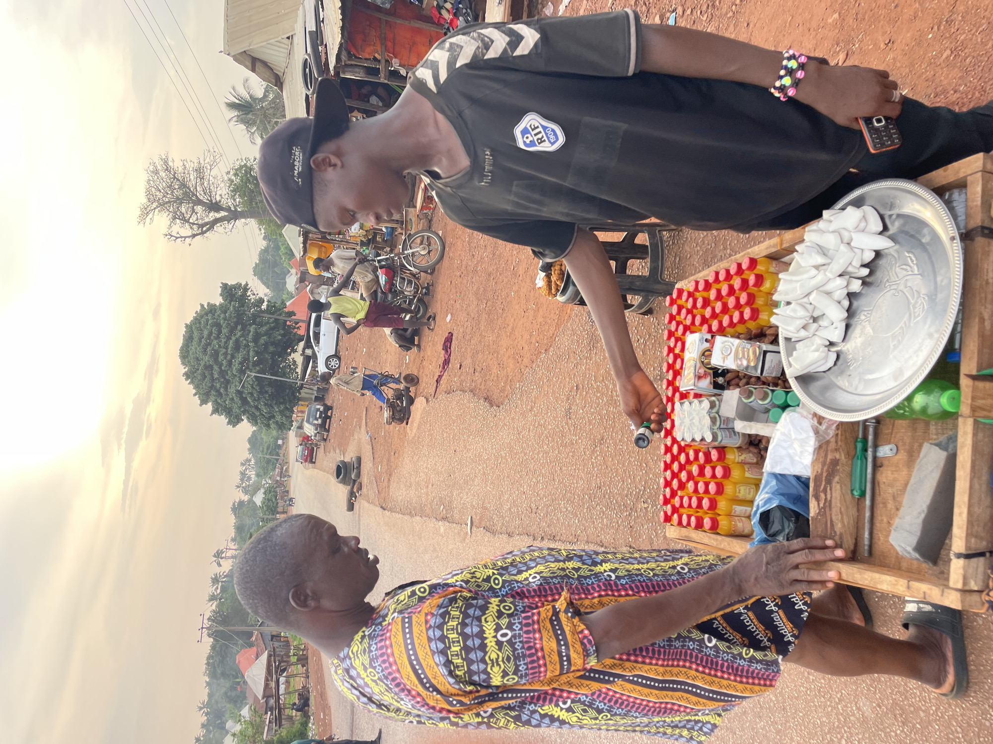 A street scene in Ogbe, Nigeria, shows two people engaged in a transaction at a roadside stall. A younger man with a black hat, black shirt, and a colorful beaded bracelet is handing a small vial to an older woman in a colorful patterned shirt. The stall is filled with bottles of juice, what appears to be coconut slices, and other items. The background shows a street with motorbikes, cars, and other people, indicating a bustling environment. The setting appears to be a typical market or commercial area in the city.