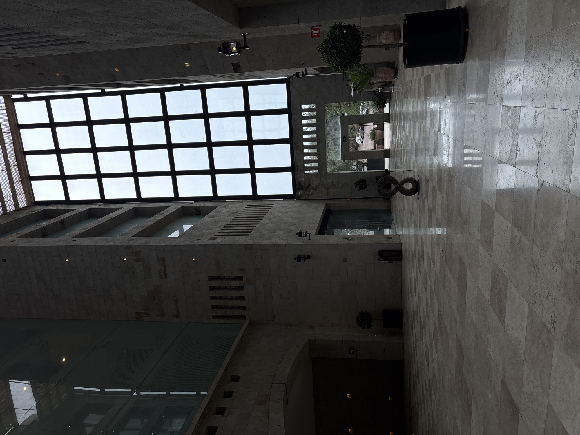 The image captures an interior space with notable architectural features. A large grid-patterned skylight is visible, allowing natural light to filter into the building. The walls are constructed from light-colored stone blocks, and a wide hallway extends into the distance, lined with potted plants and geometric art installations. The floor is tiled with a similar stone pattern. The location is Ciudad López Mateos, Mexico. There are no people visible.