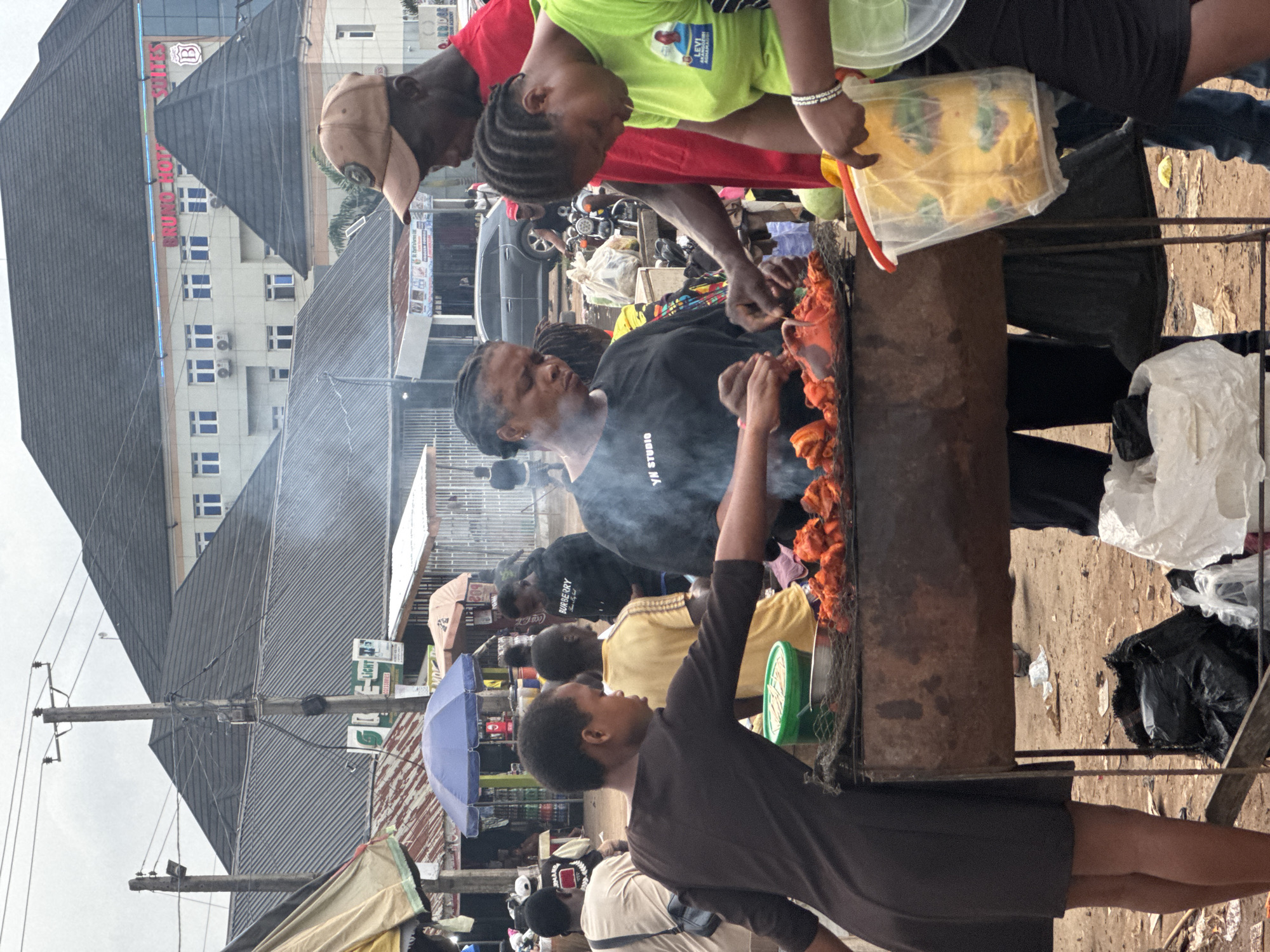 The image depicts an outdoor market scene in Obinze, Nigeria, with a focus on a food stall where chicken is being grilled. A woman in a black t-shirt with "YN STUDIO" printed on it is actively attending to the grilling chicken. To her right, a man wearing a red shirt and a tan baseball cap appears to be assisting or supervising. Another woman, wearing a bright lime green t-shirt, is holding a clear plastic container filled with yellow items, possibly food ingredients or packaging. The grill itself is a rustic, metal contraption placed on a stand. Smoke rises from the cooking chicken, creating a hazy atmosphere. In the background, various market stalls with awnings and umbrellas are visible, along with buildings, including what appears to be a hotel named "BRUNO HOTEL SUITES." A dark-colored car is parked behind the food stall, and other people are scattered throughout the scene, some blurred due to motion or distance. The ground is dirt and appears somewhat littered with discarded items, including plastic bags. The overall impression is one of a busy, active marketplace.