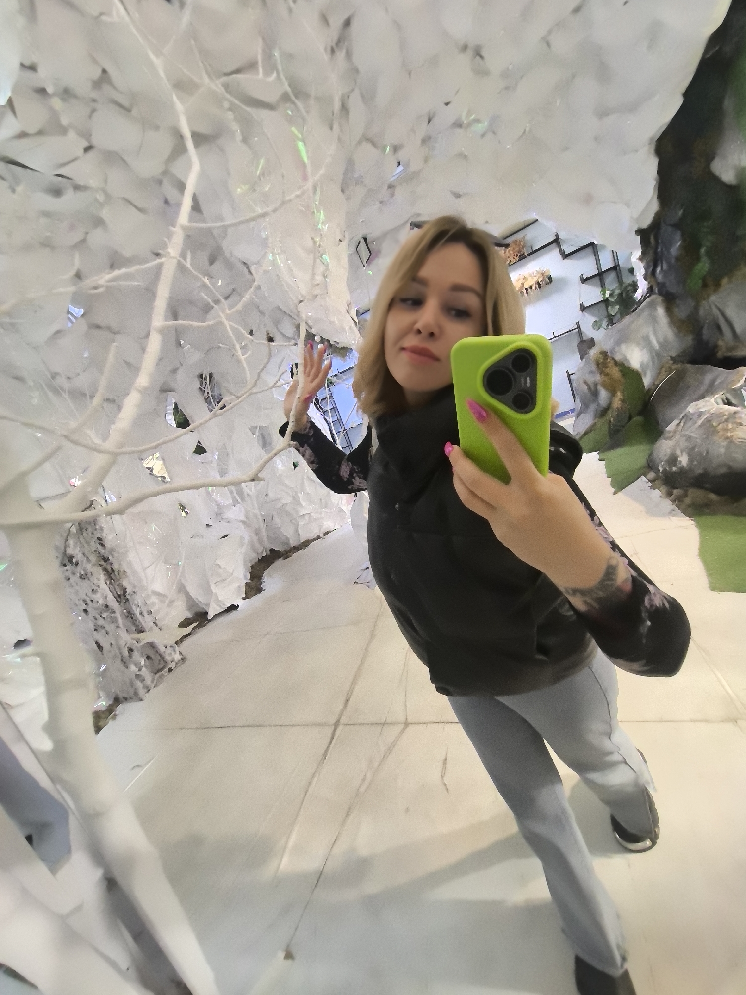 A woman with blonde hair is taking a selfie with a green iPhone in what appears to be an indoor environment, possibly an exhibition or a themed space. She is wearing a black vest over a long-sleeved floral patterned top and light grey pants. Her fingernails are painted pink. The background is decorated with artificial white trees or branches, some of which have shiny or crystalline textures. In the far background, there's a glimpse of shelves and wall decorations, as well as artificial rocks and plants. Based on the details, it appears to be a decorated installation or part of a retail establishment in Izhevsk, Russia.