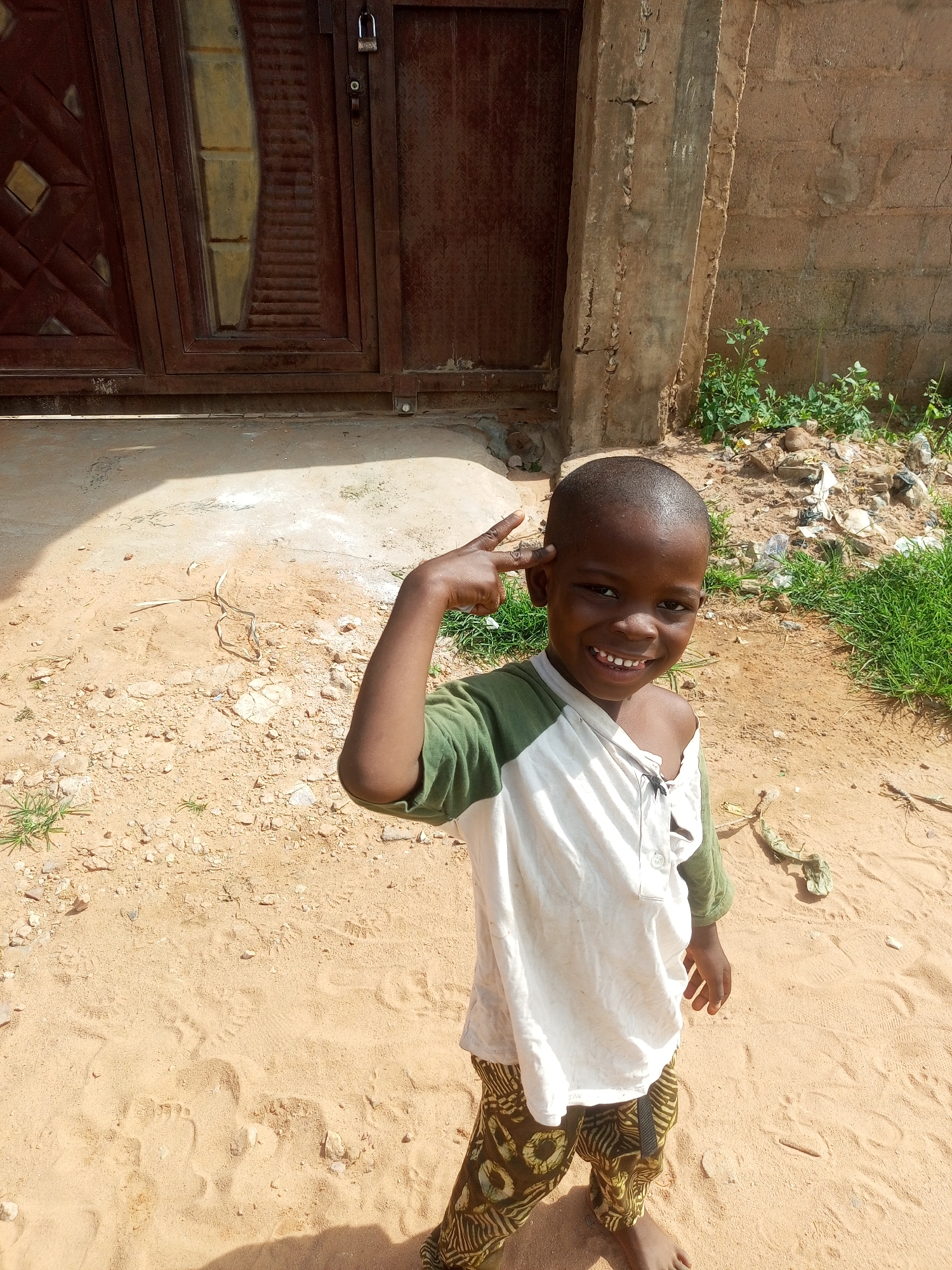 A young boy stands in the foreground on sandy ground, his head turned towards the camera. He has a broad smile, revealing his teeth. His right hand is raised to his head, with his index and middle fingers extended in a peace sign. He is wearing a green and white long-sleeved shirt and patterned pants. Behind him, to the left, is a large, dark brown metal gate with a lock. To the right, a concrete pillar stands next to a brick wall. The ground is a mixture of sand, small stones, and dry debris, with some patches of green grass and weeds growing near the wall. The scene appears to be outdoors, likely in a residential area.
