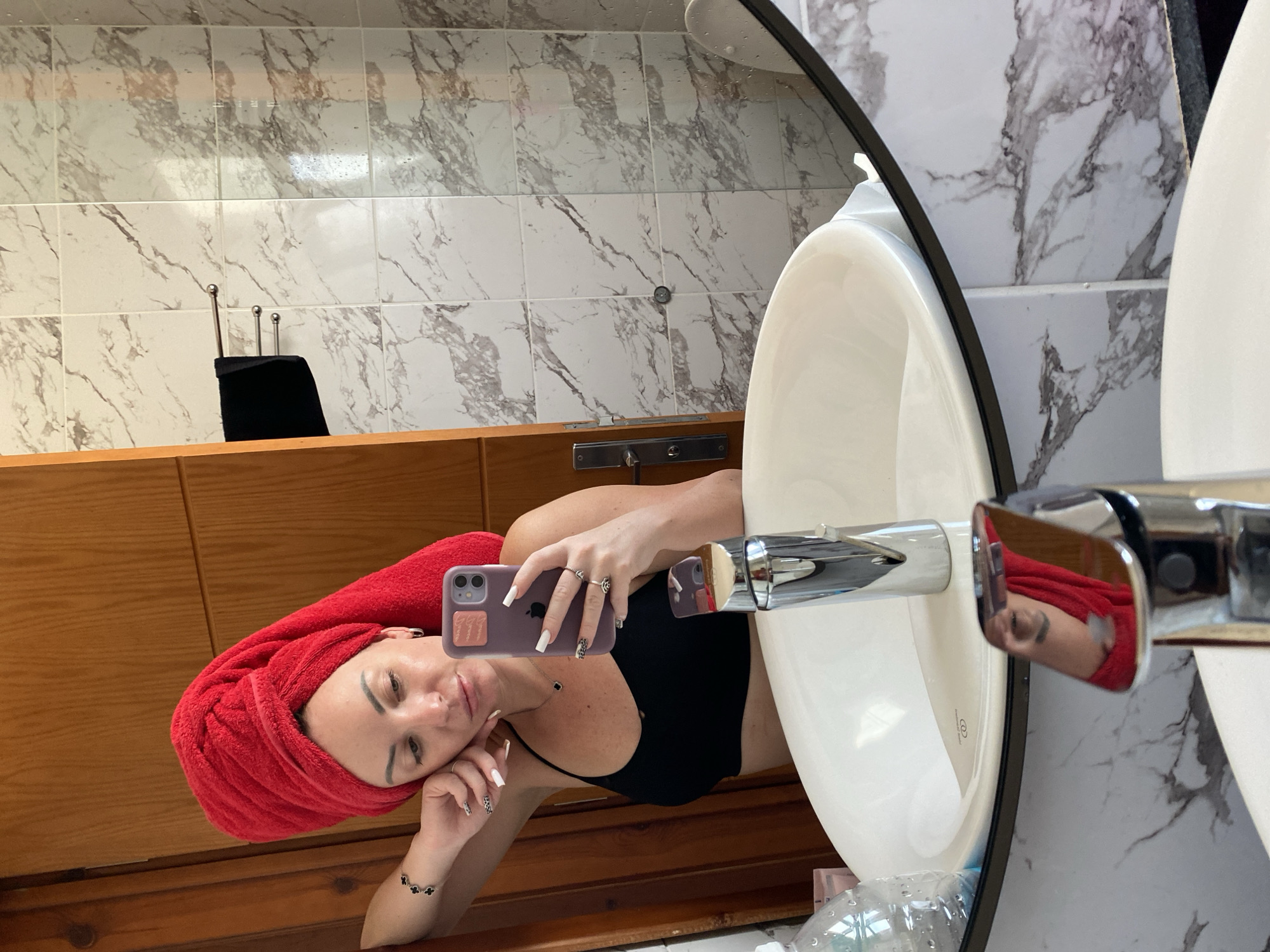 A woman with a red towel wrapped around her head and a black sports bra is taking a selfie in a bathroom mirror. She is wearing a necklace and several rings on her fingers. The bathroom features marble-patterned tiles and a wooden door. A sink with a chrome faucet is visible in the foreground. The overall setting suggests a hotel bathroom, possibly in Sharm El Sheikh, Egypt, given the context of the location.