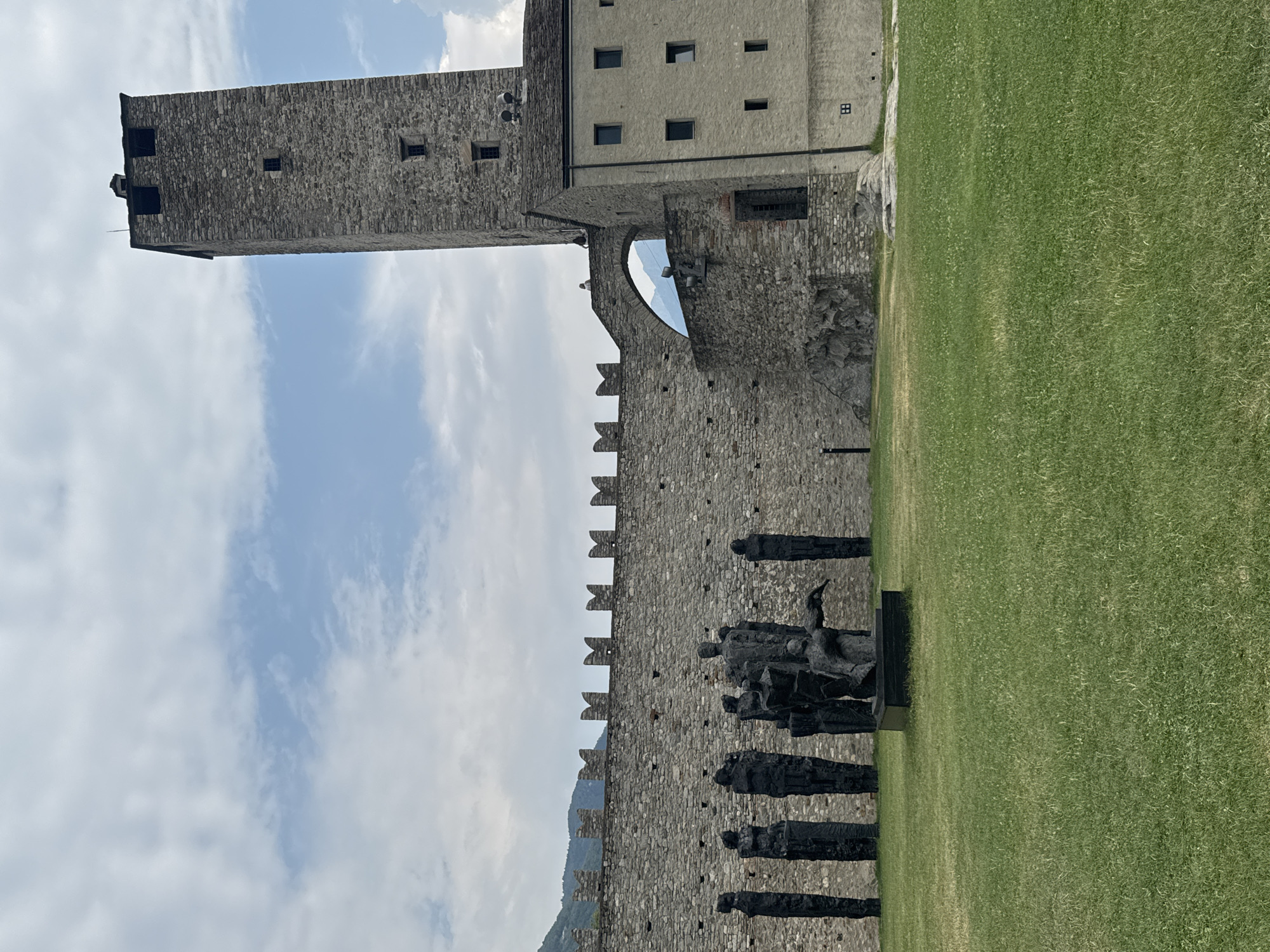 The image depicts the Castel Grande in Bellinzona, Switzerland. In the foreground, a meticulously manicured green lawn is visible, with a group of dark, abstract human-like sculptures arranged on it. These sculptures appear to be made of metal and vary in height and form, with one central sculpture depicting multiple figures in a dynamic pose. Behind the lawn and sculptures, a thick, weathered stone wall stretches across the scene, topped with crenellations. To the right of the wall, a tall, imposing stone tower rises into a partly cloudy sky. Adjacent to the tower is a lighter-colored stone building with multiple windows. An arched stone walkway connects the wall to the tower, and a single person can be seen standing on this walkway. The background features rolling hills covered in vegetation, suggesting a mountainous landscape. The sky is a mix of blue sky and white clouds.