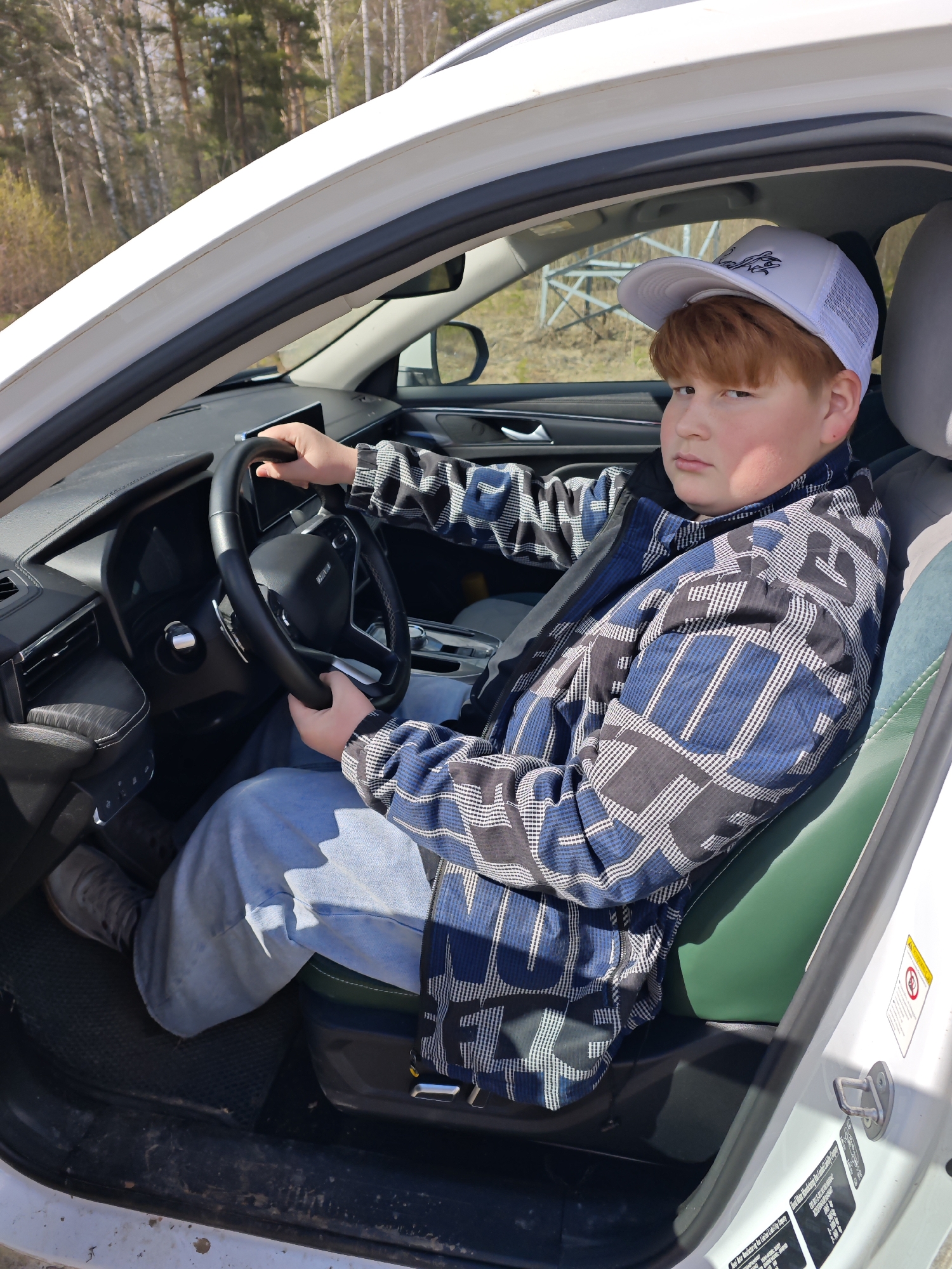 A ginger-haired boy wearing a white baseball cap, a blue, black, and white patterned jacket, and jeans sits behind the steering wheel of a white car. He is wearing a seatbelt. The car’s interior features a black dashboard and steering wheel, green leather seats, and gray headrests. Outside the car, trees and a metal structure are visible in the background. The location is likely Chaikovsky, Russia.