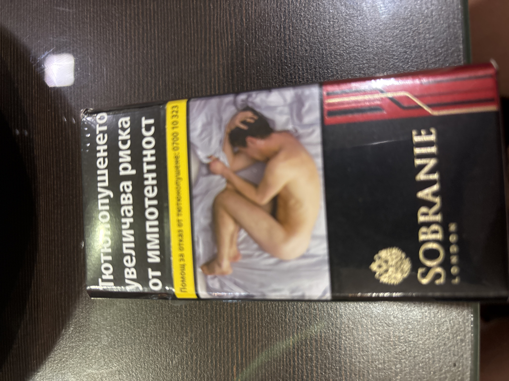 The image displays a pack of "Sobranie" cigarettes. The front of the pack features text in Bulgarian stating that smoking increases the risk of impotence, followed by a helpline number. Below this text, there is a graphic image of a naked person lying in bed. The bottom of the pack shows the "Sobranie London" logo and a red and gold striped design. The pack is positioned on a dark, textured surface, possibly a table. The background is out of focus but appears to be a dark, wooden surface with some faint reflections. There is no discernible information in the image that would place it in the city of Montana, Bulgaria.