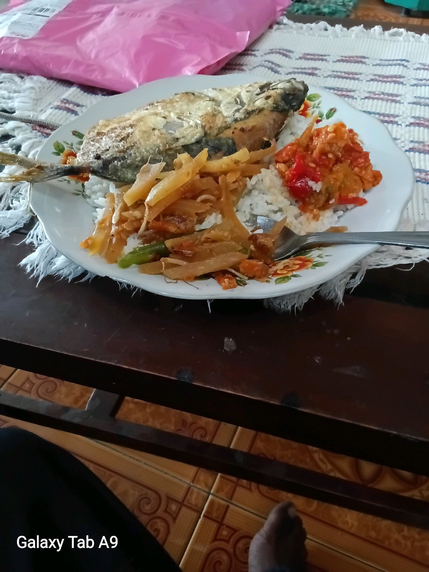 A fried fish sits atop a plate of white rice, accompanied by a side of stir-fried vegetables and a spoonful of red chili paste. The plate, adorned with floral patterns, rests on a textured placemat. In the background, a pink plastic bag is visible, adding a splash of color to the scene. The image appears to be taken on a wooden table, with a glimpse of tiled flooring and a person's foot and leg in the lower portion of the frame. The text "Galaxy Tab A9" is overlaid on the bottom left corner, indicating the device used to capture the image. While the image does not explicitly show the city of Trenggalek, Indonesia, the presence of traditional Indonesian food and common household items suggests a domestic setting within the region.