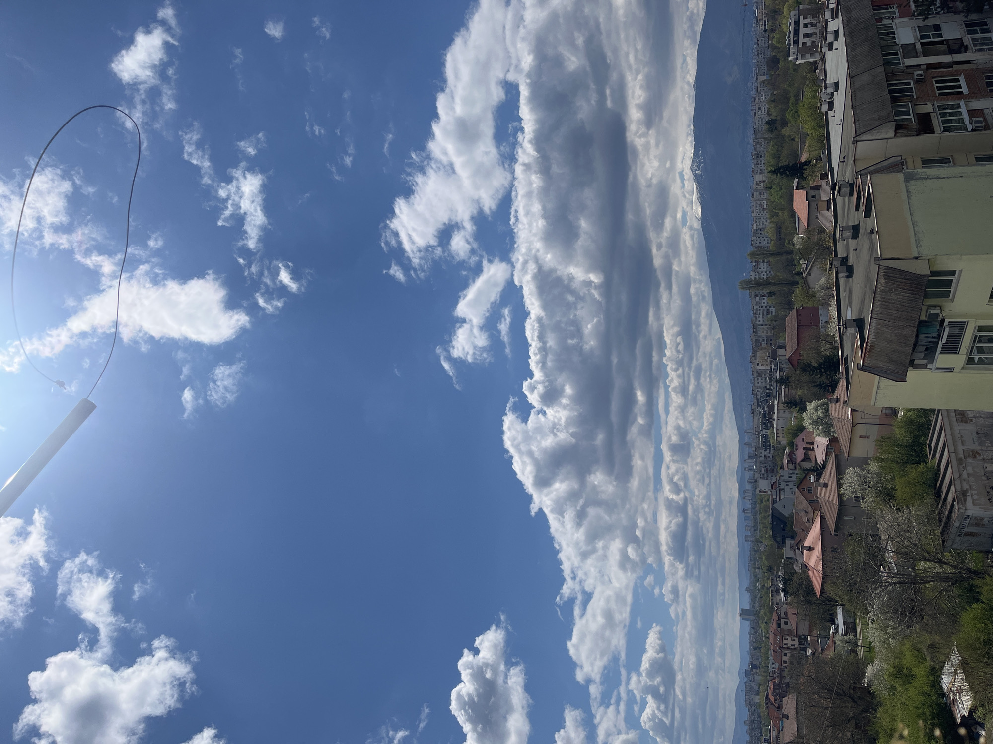 This image presents a panoramic view of Sofia, Bulgaria, capturing a dynamic urban landscape against a majestic mountainous backdrop under a bright, partly cloudy sky. The perspective appears to be from an elevated position within the city.
The sky dominates the upper portion of the frame, a vivid blue canvas adorned with numerous white, puffy cumulus clouds. Some clouds are scattered, while others coalesce into a more continuous, elongated band, particularly noticeable above the distant mountains. A strong sun flare emanates from the upper left, indicating bright daylight, likely during the morning or afternoon. A thin, dark cable or wire forms a large, graceful loop in the upper left quadrant of the sky, and a white pole extends from the lower left corner towards the sun, possibly part of a railing or antenna structure.
The city sprawls across the midground and foreground, characterized by a mix of residential buildings. Many structures feature traditional red or orange tiled pitched roofs, interspersed with taller apartment blocks, some with flat, grey rooftops. Details such as windows and air conditioning units are visible on building facades. Lush greenery, including trees and bushes, some with white blossoms, is abundant throughout the urban area, suggesting a spring setting.
In the background, a prominent mountain range stretches across the entire horizon. Patches of snow are visible on the mountain peaks, confirming the season as likely early to mid-spring.
No specific human activity is depicted, making it a static landscape scene. While some faded markings might be present on a building face to the far right, no clear or legible text is discernable anywhere in the image. The overall impression is one of a clear, pleasant day over a vibrant city situated at the base of impressive mountains.