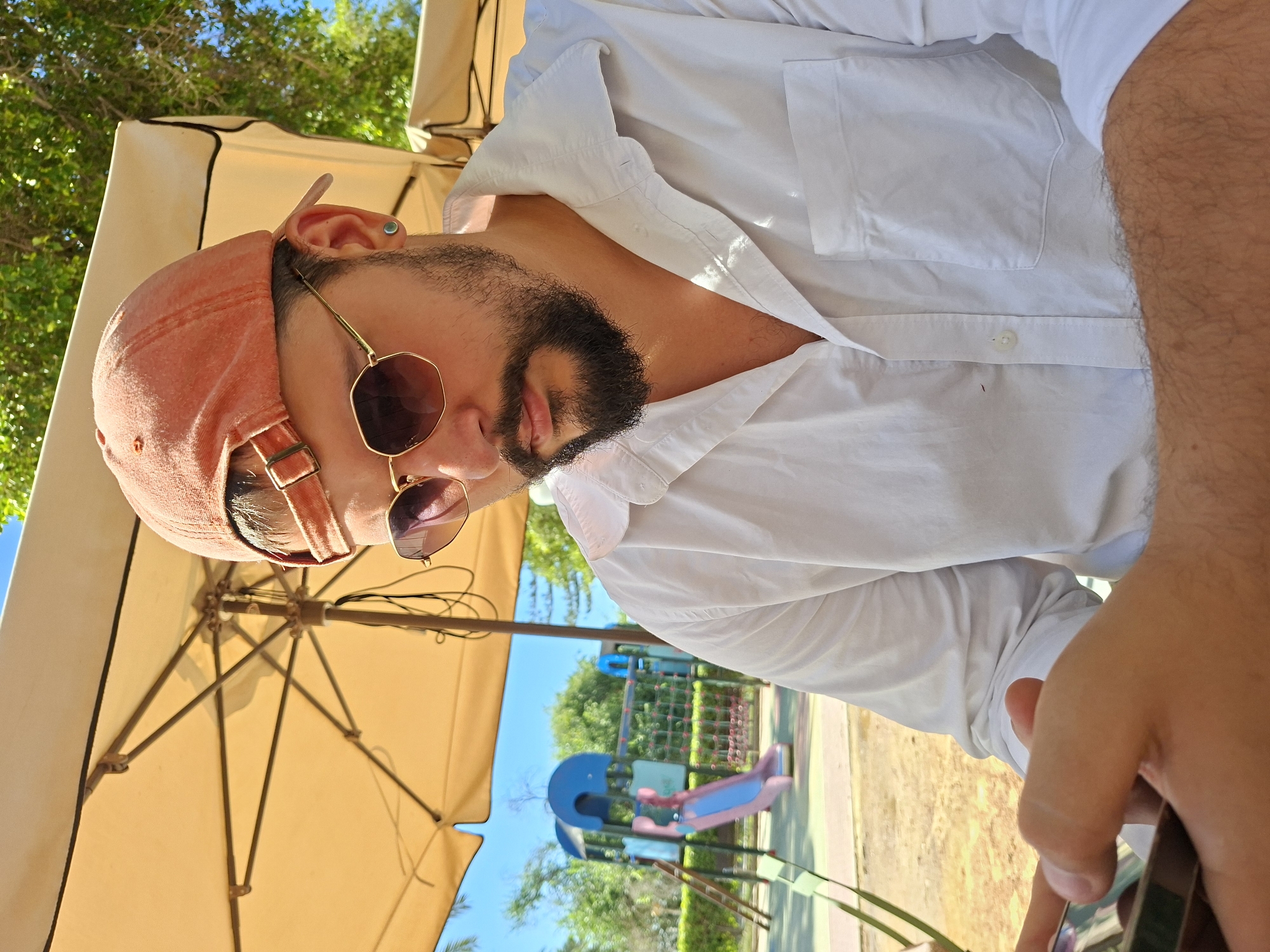 A man with a beard and a pink baseball cap is sitting outdoors, holding a smartphone and looking down at it. He is wearing a white button-down shirt and sunglasses. Behind him, a children's playground with blue and pink structures is visible, along with a yellow umbrella providing shade. The setting appears to be a park or outdoor seating area in Alicante, Spain, given the bright sunlight and Mediterranean foliage.