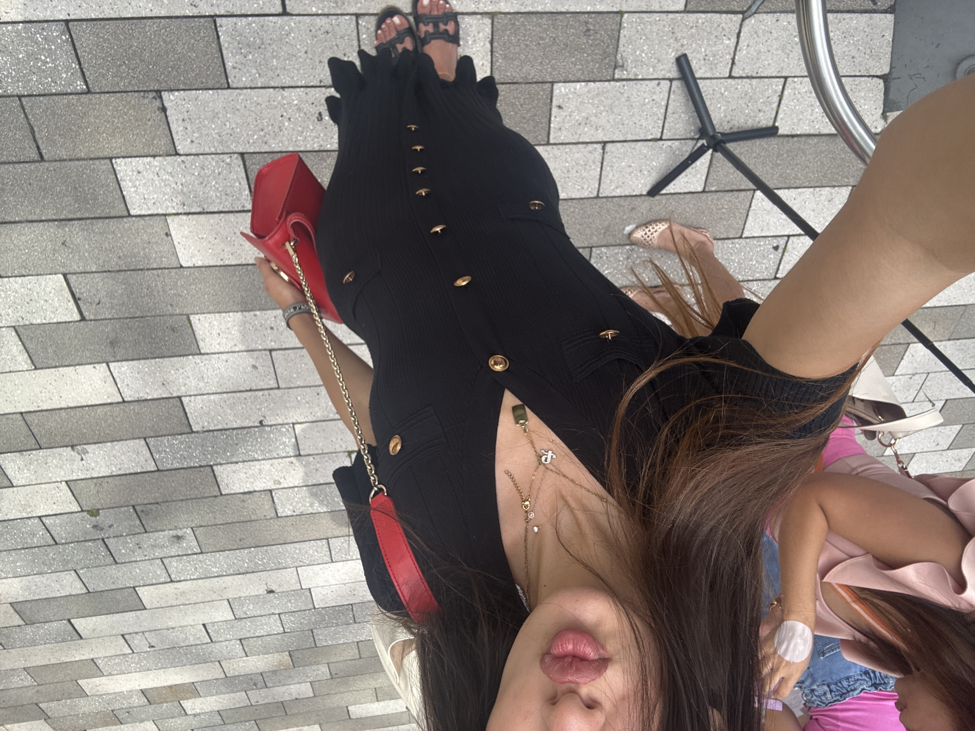 The image depicts a person's upper body and legs, taken from a high angle, outdoors on a paved surface. The individual is wearing a black, form-fitting dress with gold buttons and black sandals. A bright red handbag with a gold chain strap is slung over the shoulder. The person has long, dark brown hair and is wearing multiple necklaces. In the background, other individuals are partially visible, also dressed in casual attire. The environment appears to be a pedestrian walkway or plaza, possibly in a commercial area, indicated by the paving stones and some outdoor furniture, including a black tripod stand and a portion of a metal railing. The lighting suggests it is daytime. The location is identified as Aventura, United States of America.
