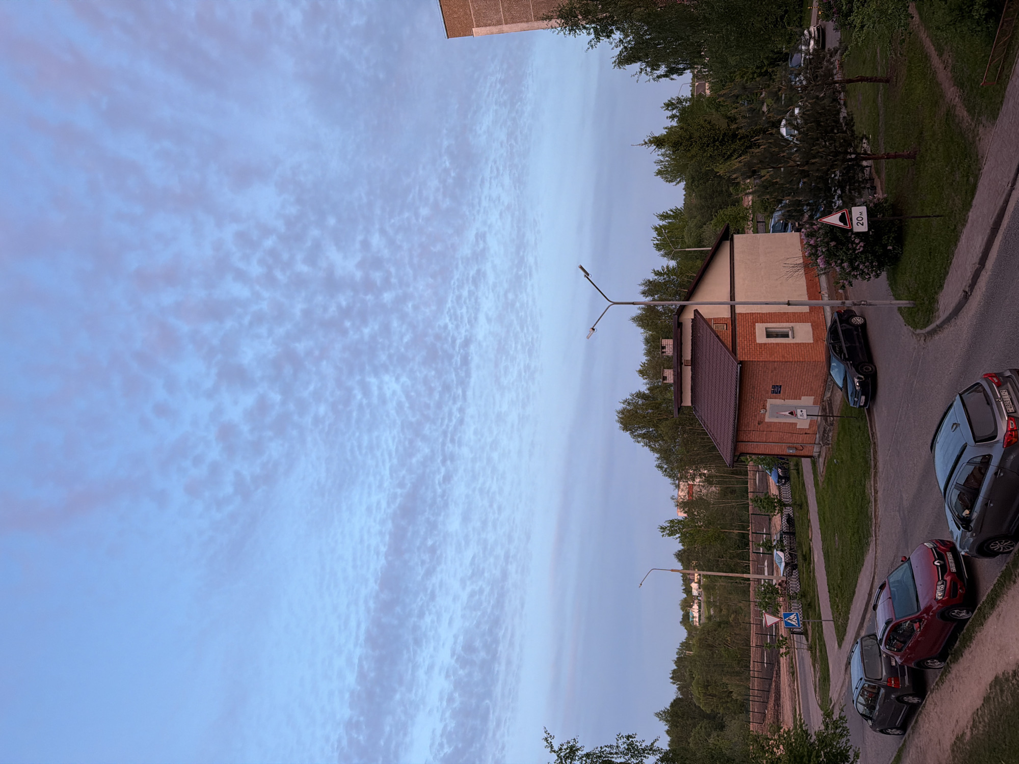 A low-angle view of a street in Unknown, Unknown shows a residential neighborhood at twilight. The sky is predominantly a pale blue with textured, cloud-like formations covering a large portion of the upper-left quadrant. Adjacent to the sky, a building with a brown roof and brick facade is visible, positioned in the middle of the image. Streetlights with a Y-shaped configuration extend from the building. A curb and sidewalk edge the lower-right quadrant, where multiple cars are parked. Trees and greenery are situated along the border between the buildings and the sky, with foliage also present in the foreground. A road sign is partially visible in the middle ground, adjacent to one of the buildings.