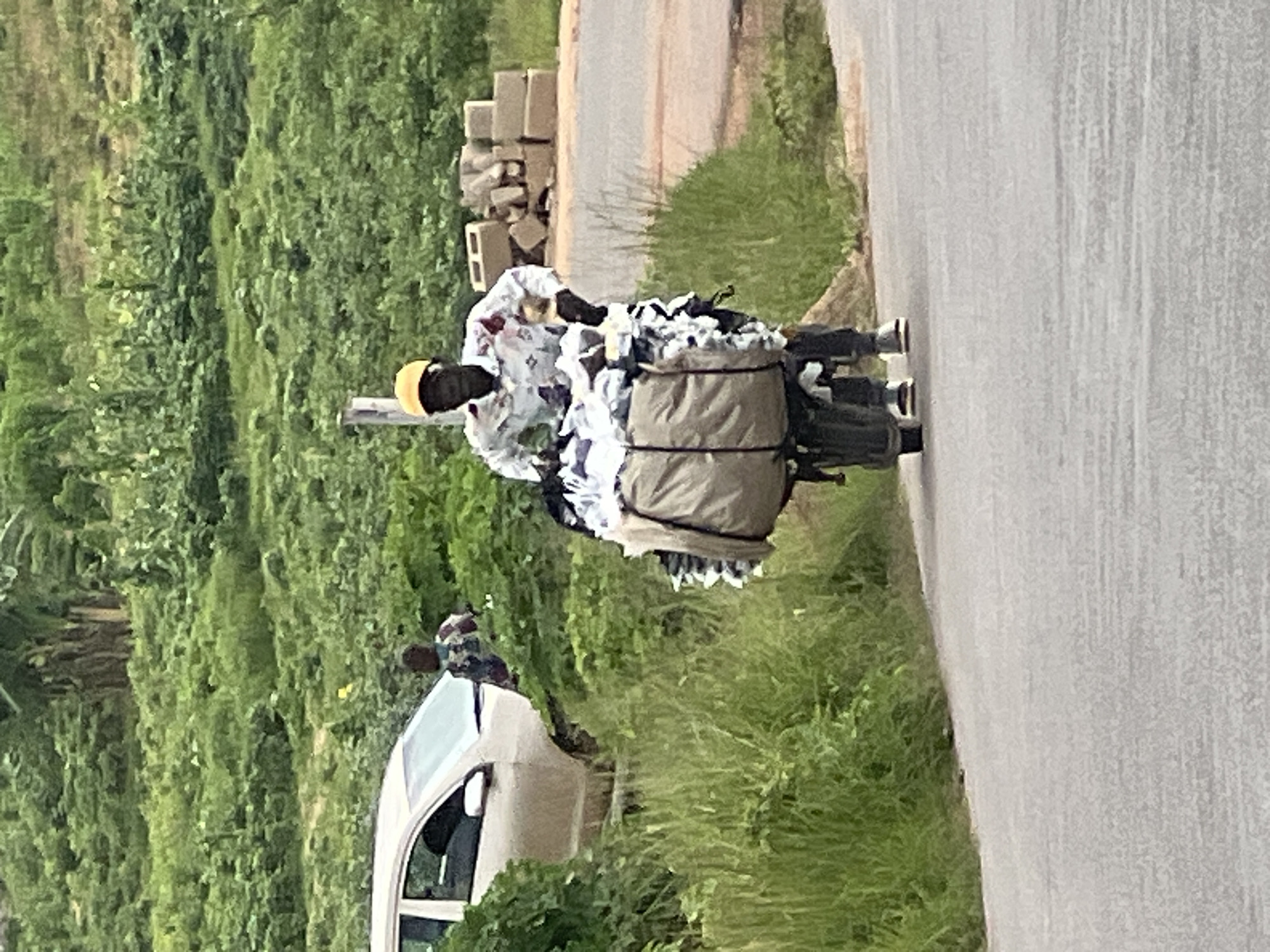 A person wearing a yellow cap and a patterned shirt is pushing a cart laden with a large, beige-colored bundle covered in fabric and clothing. The cart has two visible wheels and appears to be a bicycle frame with an attached cargo area. The person is walking on an asphalt road. To the left of the road, there is tall green grass and bushes, and further back, a light-colored car is parked. Behind the car and grass, a hill is covered in dense green vegetation. In the background, to the right of the person, is a stack of beige cinder blocks. The overall environment suggests a rural or semi-urban setting with abundant greenery. The image was taken in Jos, Nigeria.