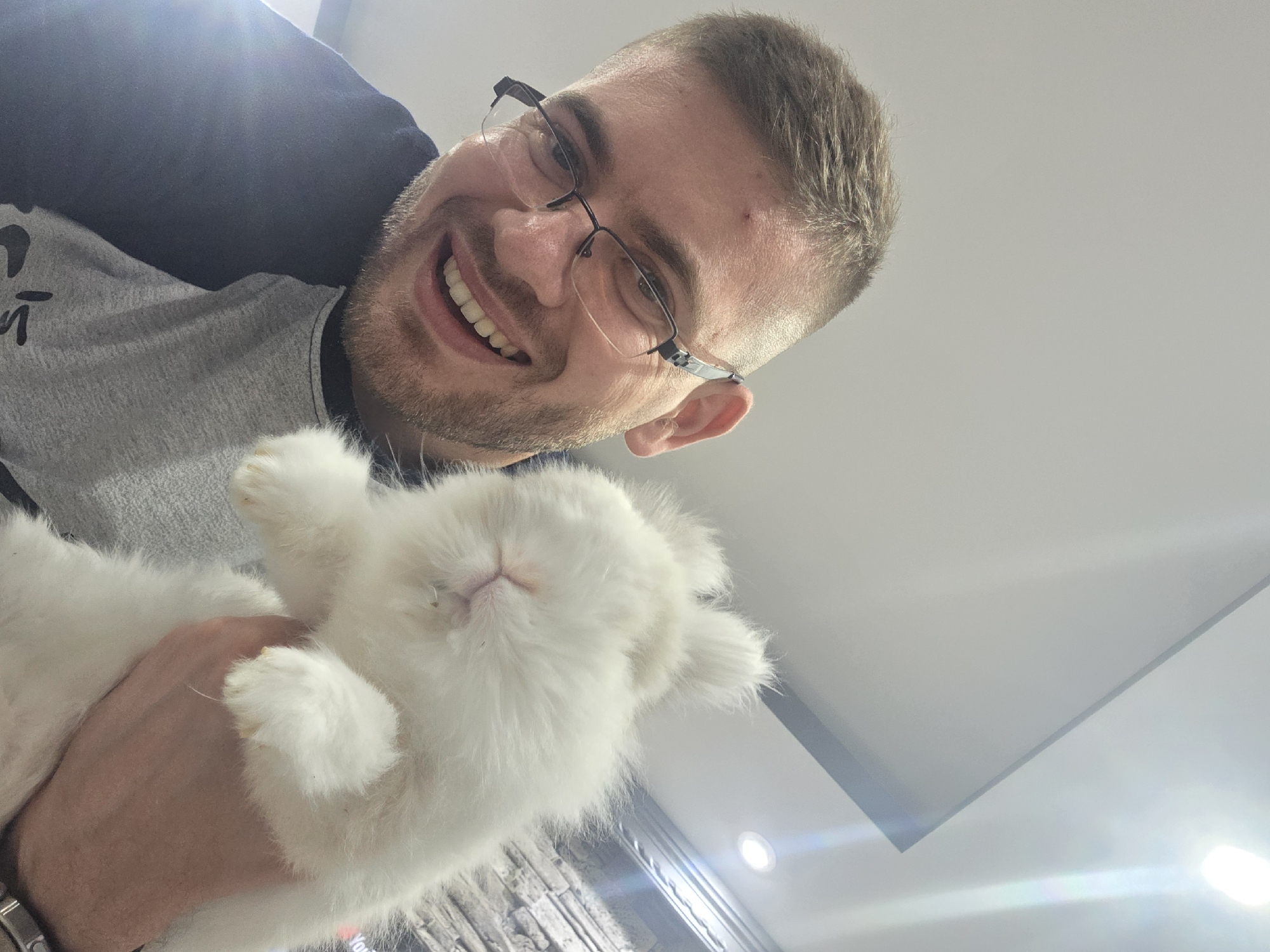 A man with short brown hair and glasses is smiling at the camera while holding a fluffy white rabbit. The man is wearing a grey and dark blue shirt. The background appears to be an interior space with a white ceiling and a stone wall visible in the lower left corner. The location within Yagodovo, Bulgaria is not discernible from the image.