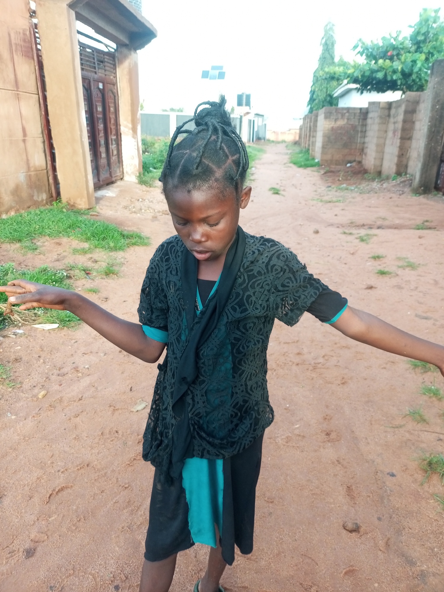 A young Black girl with braided hair is standing on a dirt road in what appears to be a residential area in Tempure, Nigeria. She is wearing a black lacy top over a teal dress, and black pants. Her right hand is extended, and she is looking down at it. The background shows a light-colored building with a dark wooden door, some overgrown grass, and a fence made of stacked concrete blocks. Trees and other buildings are visible in the distance. The sky is overcast.