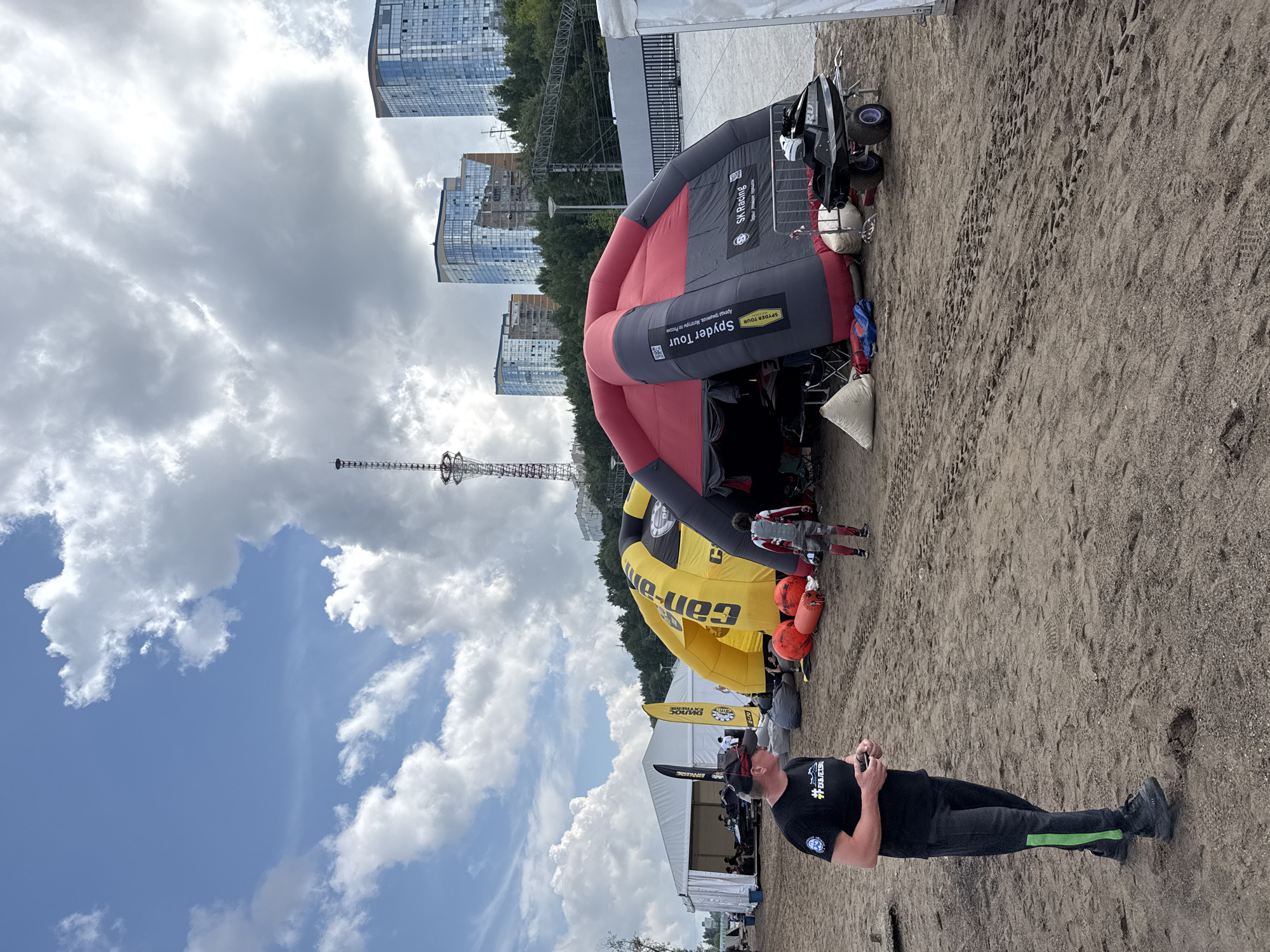 An outdoor event space is depicted in Perm, Russia, under a partially cloudy sky. The scene features a sandy ground covered in tire tracks and footprints, indicating recent activity. In the foreground, a man in a black t-shirt and dark pants with a green stripe on the leg is standing, holding a phone. Behind him, two large inflatable structures are prominent: a yellow and black "Can-Am" branded inflatable and a red and black "Spyder Tour" branded inflatable. Several large, orange inflatable buoys are placed near the yellow structure. A person in a red and grey racing suit is walking away from the red and black inflatable. A personal watercraft is parked on a trailer to the right of the red and black inflatable. In the background, a tall radio tower stands against the sky, and modern glass-fronted buildings rise above a treeline. A white tent is visible on the far left of the image, with people seated inside. The overall impression is of an organized event, possibly related to water sports or racing, set up in an urban environment.
