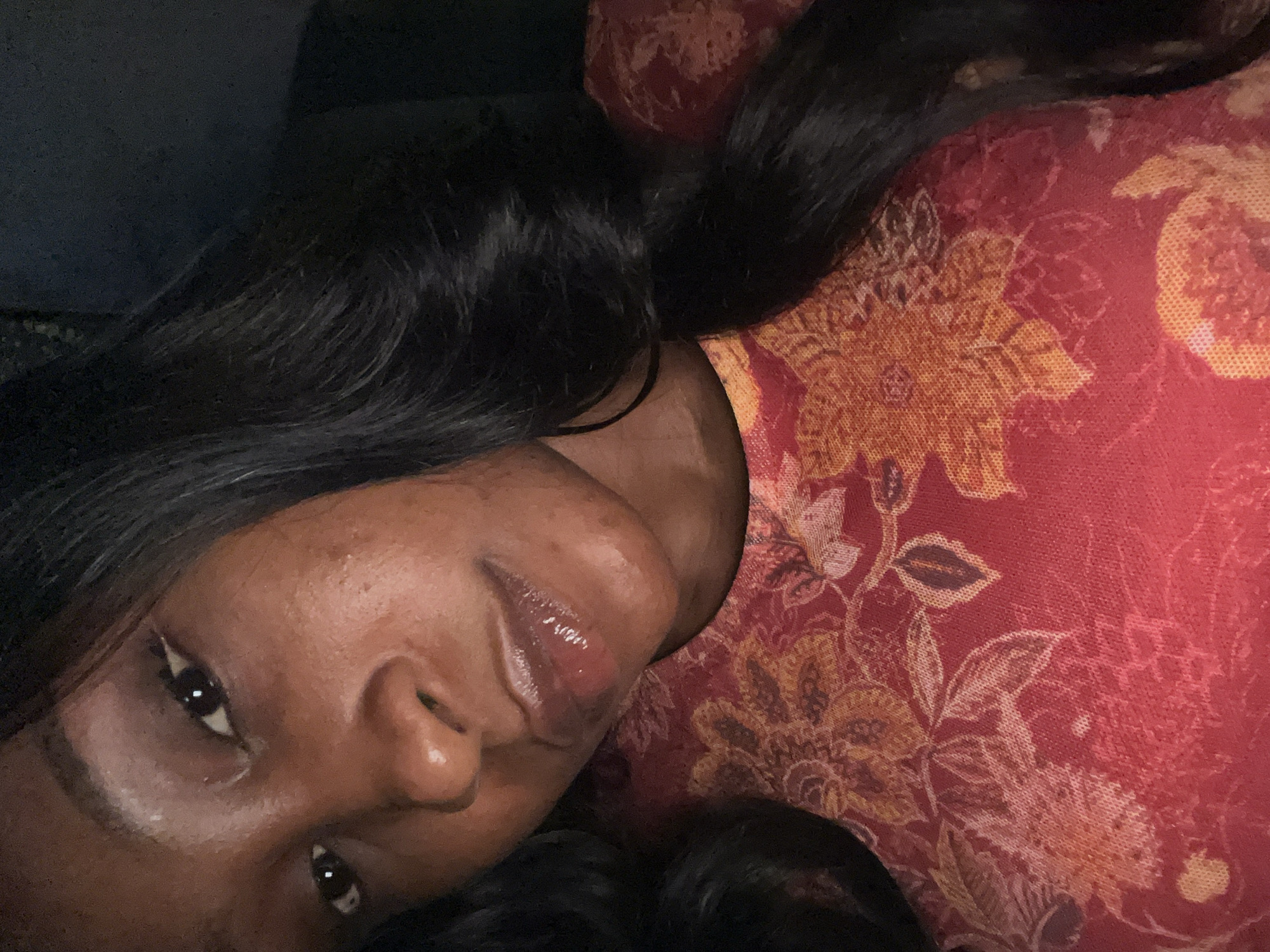 A close-up selfie shows a young Black woman with long, dark hair and fair skin against a dark background. She is wearing a red and orange floral print top. The lighting in the image is dim, and the focus is on her face. The image does not provide any information about the location in Abuja, Nigeria.