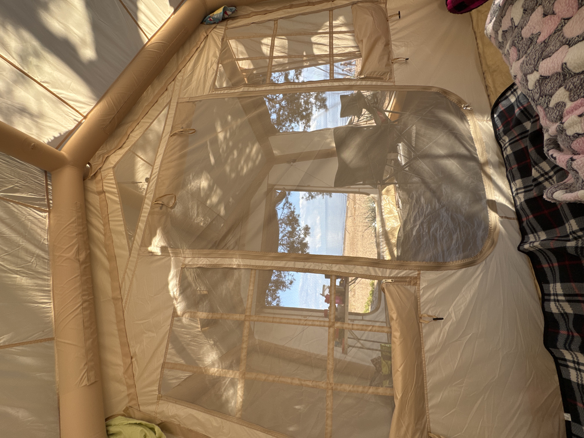 The image provides an interior view of a large, tan-colored inflatable tent, possibly located near the coast in Zelenaya Roshcha, Russia. Sunlight streams through the tent's mesh windows, casting dappled shadows from overhead trees onto the fabric. Outside the tent, a sandy beach and the calm blue sea are visible under a bright sky.
Inside, the tent is furnished with camping chairs and a portable table laden with various items, including containers and possibly food or drinks. Two camping chairs are set up, one in the foreground and another further back, both facing outwards towards the view. The floor of the tent is covered with blankets, one a plaid pattern and another a soft, patterned fleece in pink and gray. The tent's structure is supported by inflatable poles, visible along the ceiling. In the distance, two figures can be seen walking along the shoreline. The overall scene suggests a relaxed outdoor camping experience.