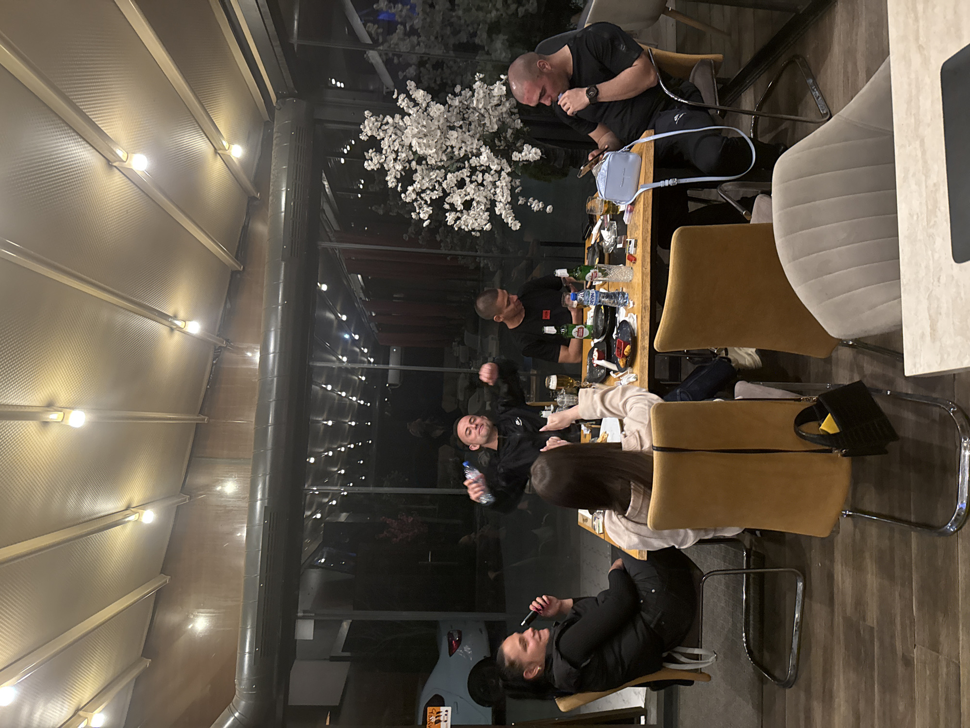 The image depicts a group of at least five individuals sitting at a table in an enclosed outdoor space, likely a restaurant or cafe. The setting appears to be nighttime, with artificial lighting illuminating the area. A white flowering tree or decoration is visible behind the glass enclosure.
The individuals are of diverse ages and genders, with most dressed in dark clothing. Some are seated and interacting around a table cluttered with bottles, glasses, and plates. One person is smoking a cigarette.
The location is in an urban environment, with a partially visible white car parked outside the enclosure. The overall scene suggests a casual gathering or social event. The city is Unknown, Unknown.