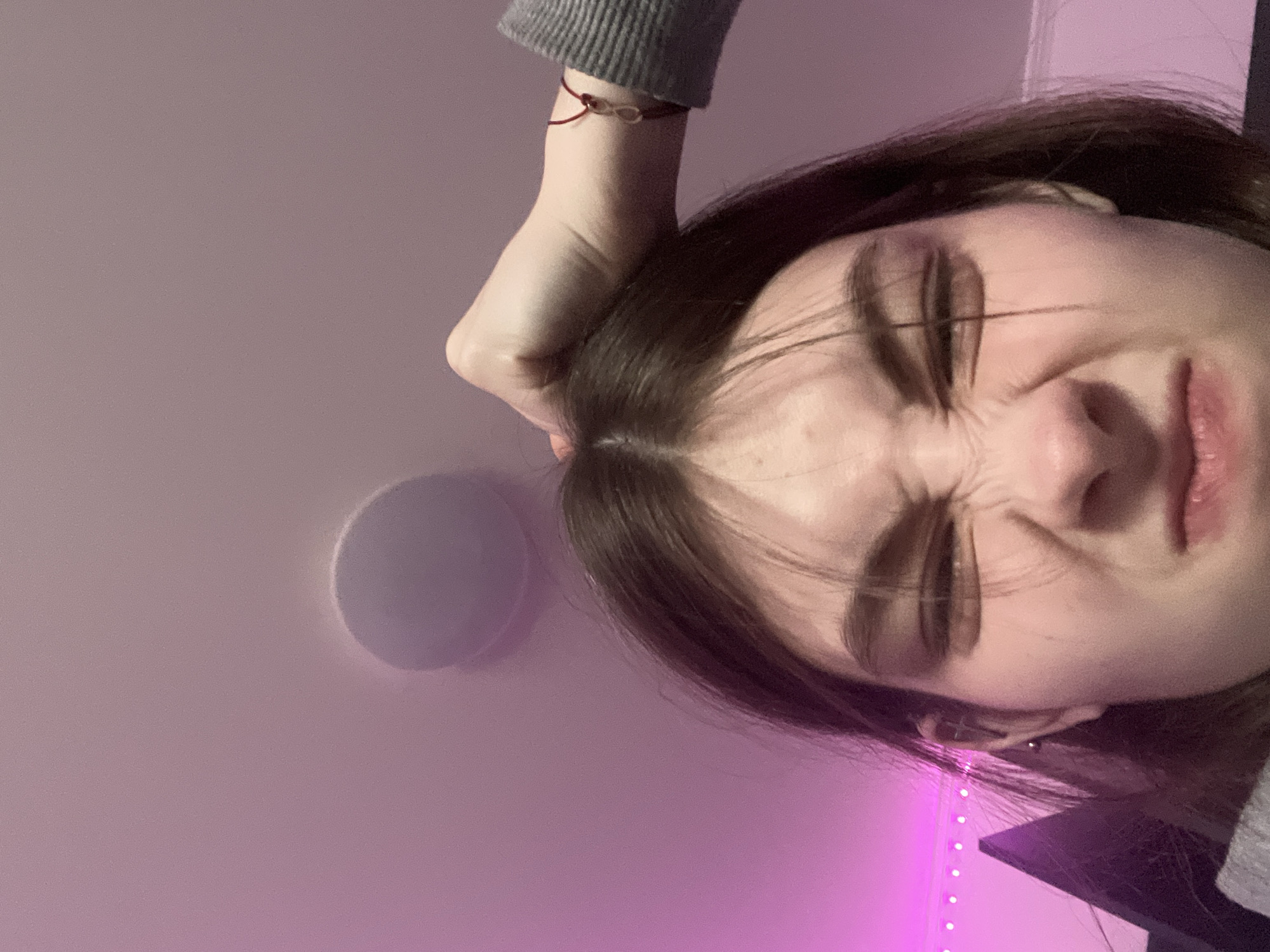 A close-up, rotated image shows a young woman with brown hair squinting her eyes shut and scrunching her nose. Her hand is positioned on her head. She has fair skin. She is wearing a gray sweater. A red string bracelet is on her wrist. An LED light strip is visible in the background. A circular object, possibly a speaker, is on the ceiling. The location is not discernible from the image and could not be identified as Saint Petersburg, Russia.