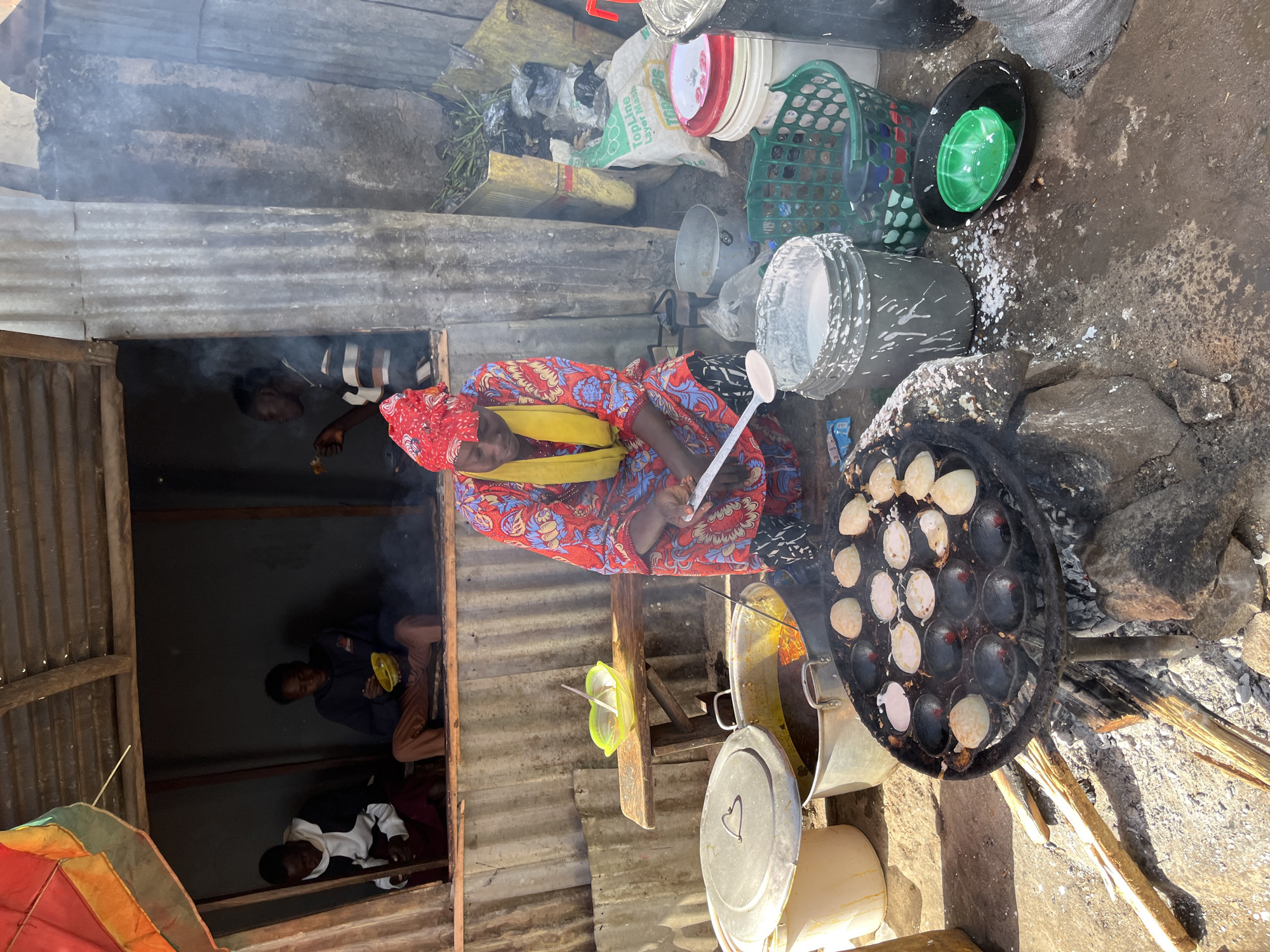 In Gura Top, Nigeria, a woman in a vibrant red and blue patterned dress and yellow headscarf is cooking small, round fritters on a charcoal stove. The cooking pan, a dark, cast-iron griddle with multiple circular molds, sits over a fire fueled by wood and rocks. Several fritters are visible in the pan, some appearing cooked and golden brown, while others are lighter in color. The woman holds a metal spatula in her right hand, poised to flip or remove the fritters. Surrounding her are various cooking implements and ingredients, including large metal pots, buckets, and a green plastic basket filled with what appears to be produce. In the background, two men sit inside a simple, open-front structure, one of whom is eating from a yellow bowl. Smoke drifts from the cooking area, adding to the hazy atmosphere of the outdoor setting. The ground is dirt and appears somewhat damp, with scattered debris and fallen leaves. The overall scene depicts a busy street-side food preparation activity.