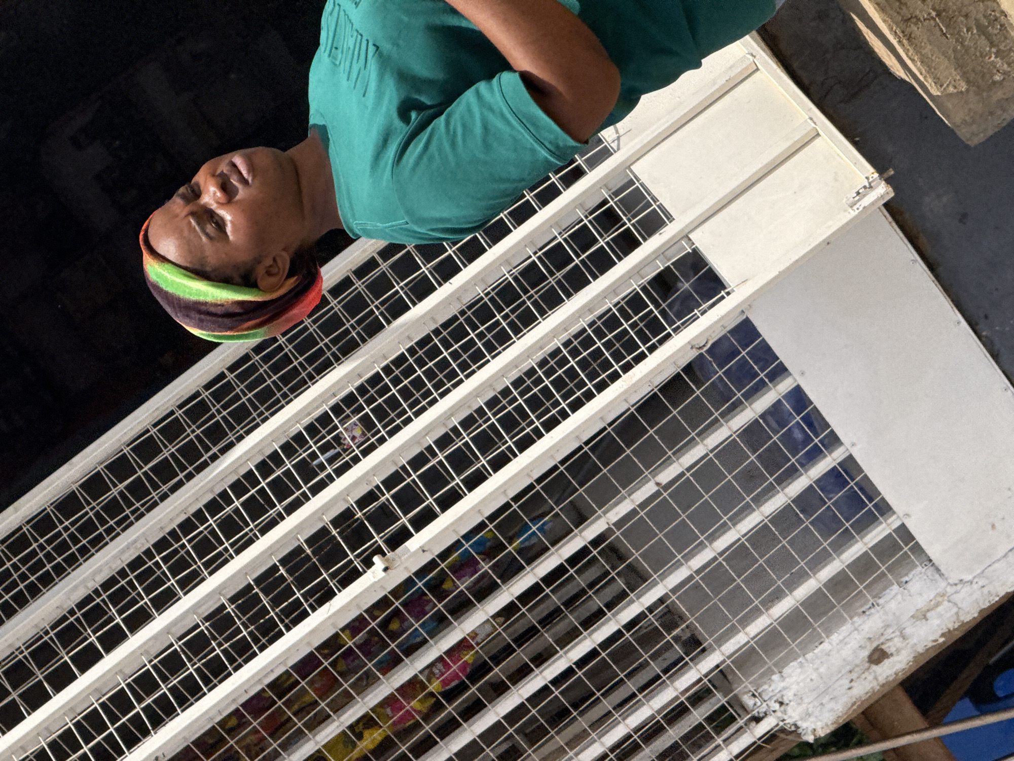 A young woman with a colorful headscarf and a teal t-shirt stands in front of a white metal gate. The gate is angled diagonally across the frame. Behind the gate, several colorful items are visible, possibly merchandise. The background is dark, suggesting the image was taken outdoors in daylight. The woman's head is tilted back and her eyes are closed, appearing to be looking upwards. Her face is illuminated by harsh sunlight, creating highlights on her forehead and cheeks. The scene appears to be in a marketplace or a shop.