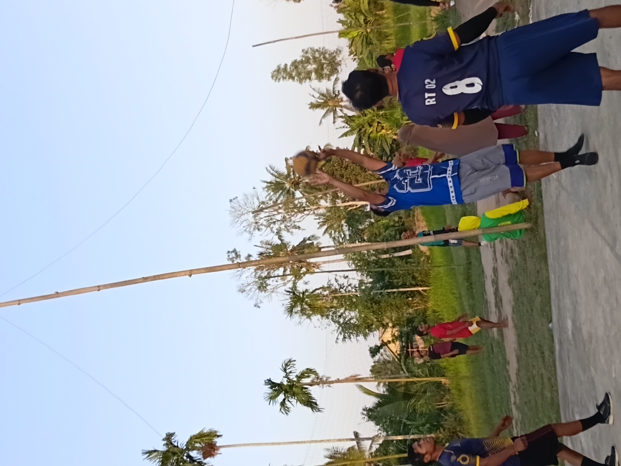 In Margo Rukun, Indonesia, on a sandy court, a volleyball game is in progress. A man in a blue and white jersey is in mid-air, about to spike a volleyball. He is wearing grey shorts with blue trim and black socks. Another man in a blue jersey with "RT 02" and the number "8" is facing away from the camera. He is wearing blue shorts. Several other people are visible in the background, including a person in a red shirt and yellow shorts, a person in a brown hijab and maroon pants, and someone in a green and yellow hooded jacket. Palm trees and other greenery surround the court, with a clear sky overhead. The game appears to be played in the late afternoon or early evening, indicated by the lighting.