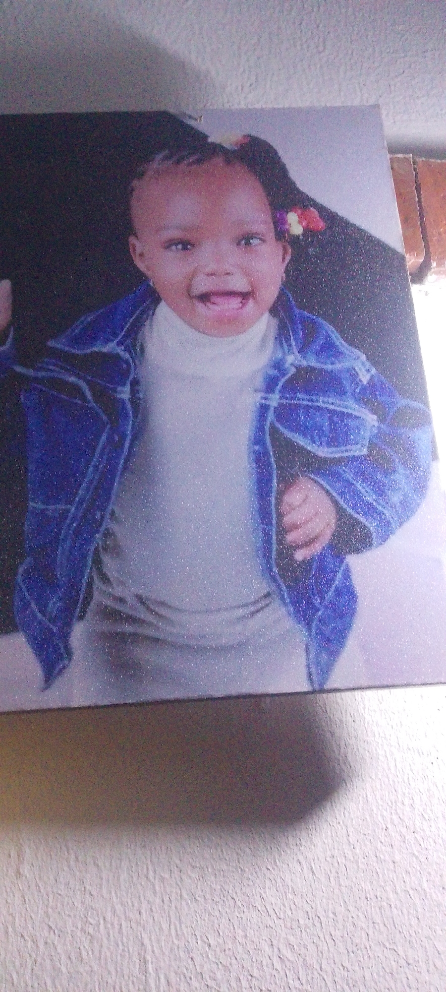 A close-up, vertical, slightly grainy photograph shows a canvas print of a young Black child. The child, likely a toddler, is smiling widely with their mouth open, and their eyes are wide and looking forward. They have braided hair with colorful hair accessories on the right side of their head. The child is wearing a white turtleneck shirt and a blue denim jacket. The background of the print is dark, with some subtle sparkles, possibly indicating a textured or metallic finish. The bottom portion of the photograph shows a textured, light-colored wall and a shadow cast on it, suggesting the print is hanging on a wall. The context of the city of Unknown, Unknown is not discernible from the image itself.