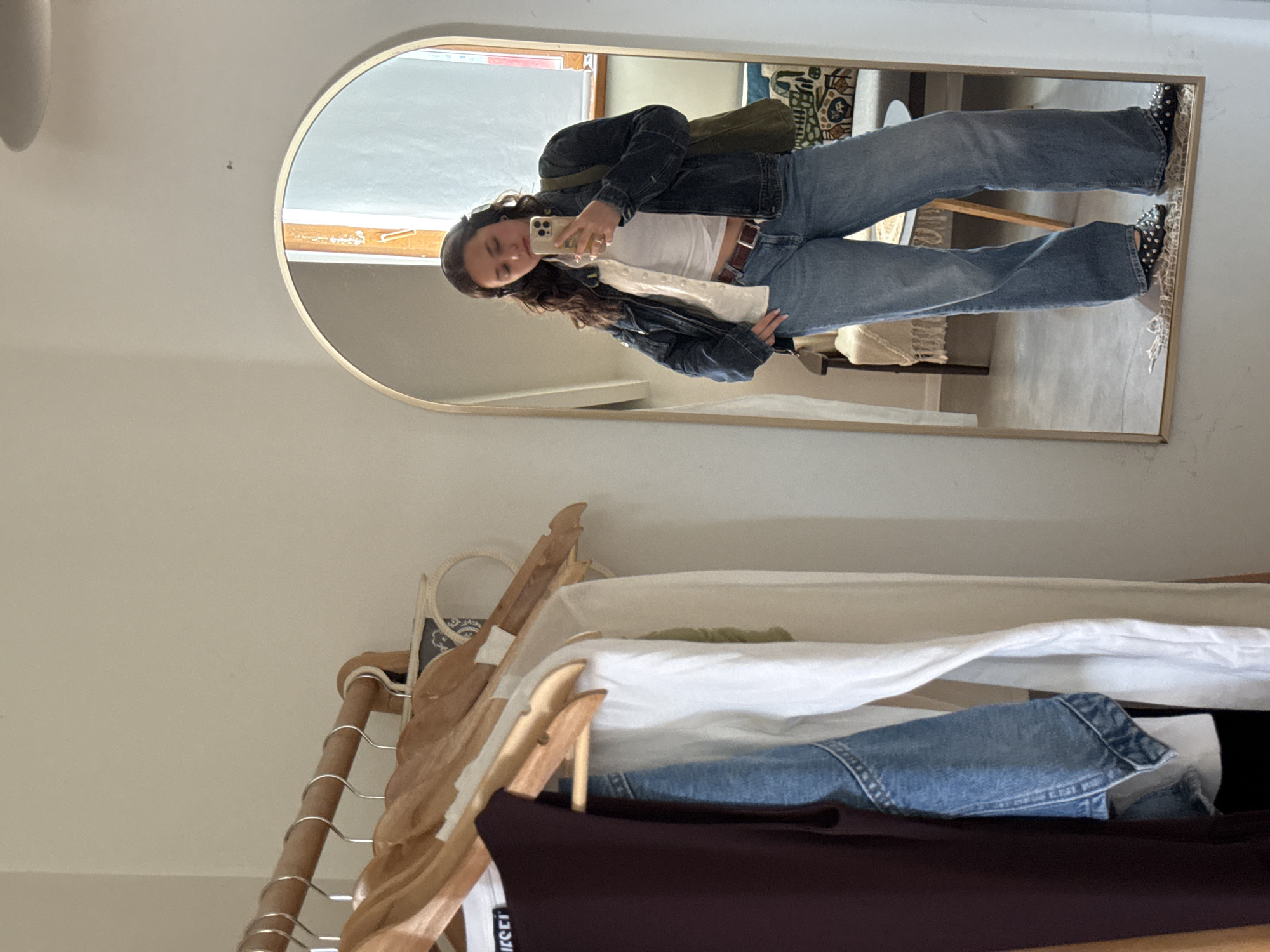 An indoor scene, possibly a dressing area or bedroom, features a young woman taking a mirror selfie. The image is oriented with the woman standing upright, reflected in a large, arched mirror with a gold frame, placed on a light-colored wall.
The woman has long, dark, wavy hair and is wearing a casual outfit consisting of a white cropped tank top, a dark denim jacket worn open, and light-wash, loose-fitting denim jeans. She has a dark brown belt with a visible buckle and black studded shoes. A dark green shoulder bag is worn across her body. She holds a smartphone to take the picture, with her head slightly tilted and a relaxed expression, suggesting contentment. Rings are visible on her fingers.
The mirror's reflection reveals an interior space with natural light coming from a window, and hints of a patterned rug and furniture. Below the mirror, a wooden clothing rack is visible, adorned with various garments on wooden hangers. These include a dark brown long-sleeved top with a "DIESEL" label visible, a white top, and light blue denim jeans. A patterned tote bag or basket also hangs on the rack.
The activity is a mirror selfie, likely to document or showcase her outfit. The scene appears to be set during the daytime, given the natural light. No specific visual cues link the image to Yerevan, Armenia, other than the provided context.