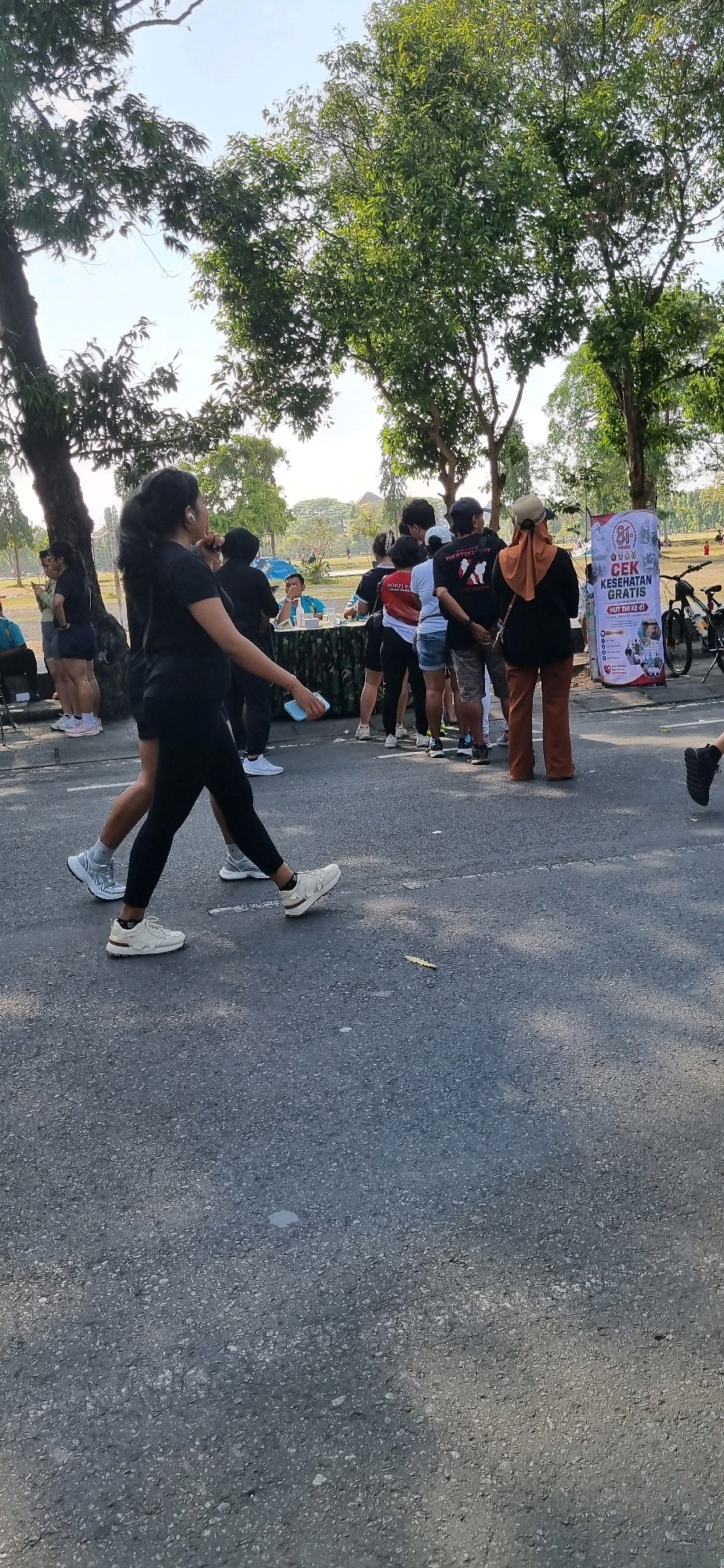 In Denpasar, Indonesia, a group of people are gathered in an outdoor public space, likely a park or street, under the shade of large trees. The scene appears to be related to a community event, possibly a health screening, as indicated by a banner that reads "CEK KESEHATAN GRATIS" (Free Health Check).
In the foreground, two women are walking on a paved road. The woman on the left, with her hair pulled back in a ponytail, is wearing a black t-shirt and black leggings, and white athletic shoes. She is holding a light blue object in her right hand and has white earbuds in her ears. The woman on the right is partially visible, wearing black leggings and white athletic shoes.
Behind the walkers, a table is set up with a patterned tablecloth, and several individuals are standing around it. A man in a teal shirt is seated behind the table, and others are queuing. Further back, a group of people are standing and observing. Among them, a woman wears an orange hijab and brown pants, and another woman has a red and white shirt.
A large banner with text and images is displayed near the right edge of the frame. Two bicycles are parked behind the banner. The environment includes lush green trees with dappled sunlight falling on the ground. In the distance, the landscape appears to be open with some structures visible. The overall atmosphere suggests a casual, organized gathering.