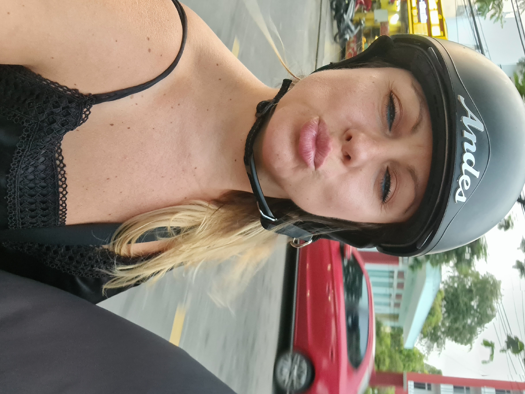 A blonde woman is featured in a close-up, selfie-style portrait, with her head tilted. She is wearing a black half-face motorcycle helmet, which has the brand name "Andes" written in white cursive script on its side. Her lips are puckered in a playful "kissy face" expression, and her light blue or grey eyes are looking towards the camera. She is wearing a black top with thin straps and intricate lace detailing along the neckline. Her blonde hair is visible under the helmet.
The background is an out-of-focus urban environment. A red car is prominently visible behind her, suggesting a street or parking area. Faint yellow road markings can be seen on the asphalt surface. Distant buildings, trees, and other vehicles (possibly motorbikes) are discernible in the soft, diffused daylight, suggesting it is daytime. The scene evokes a casual moment in Đà Nẵng, Vietnam.