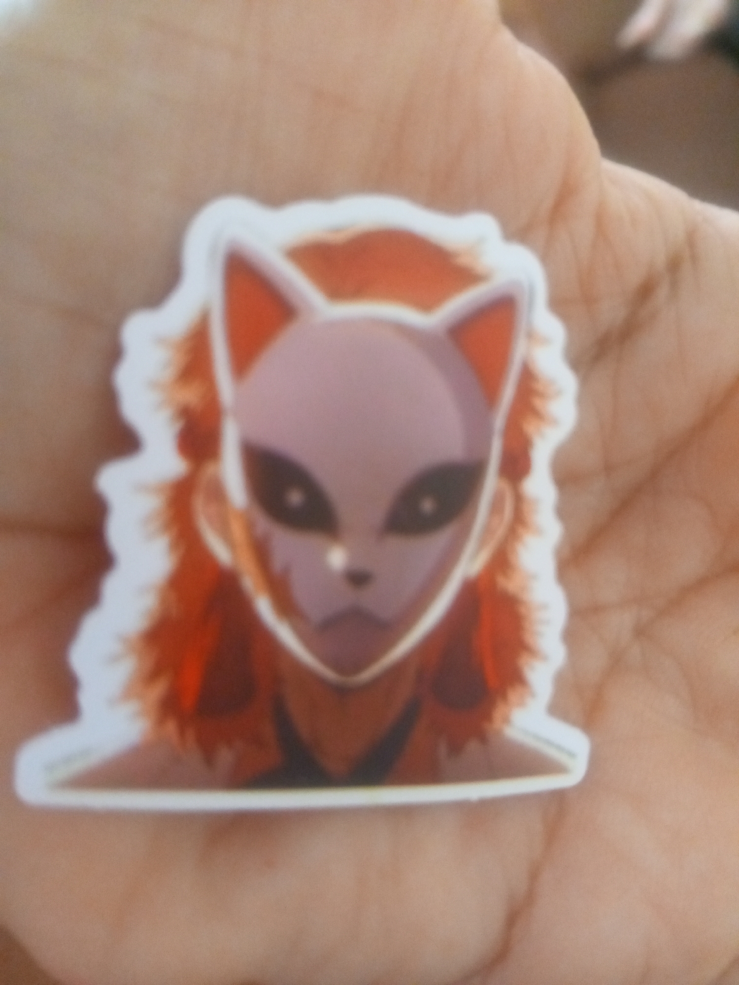 A sticker of an anime character wearing a fox mask is held in a person's palm. The sticker features the character's face with a light purple mask that has black eyes and a small nose and mouth. The character has reddish-brown hair and a white border around the sticker. The background is blurry, showing the lines and texture of a person's hand. There is no discernible location in Altagracia, Venezuela.