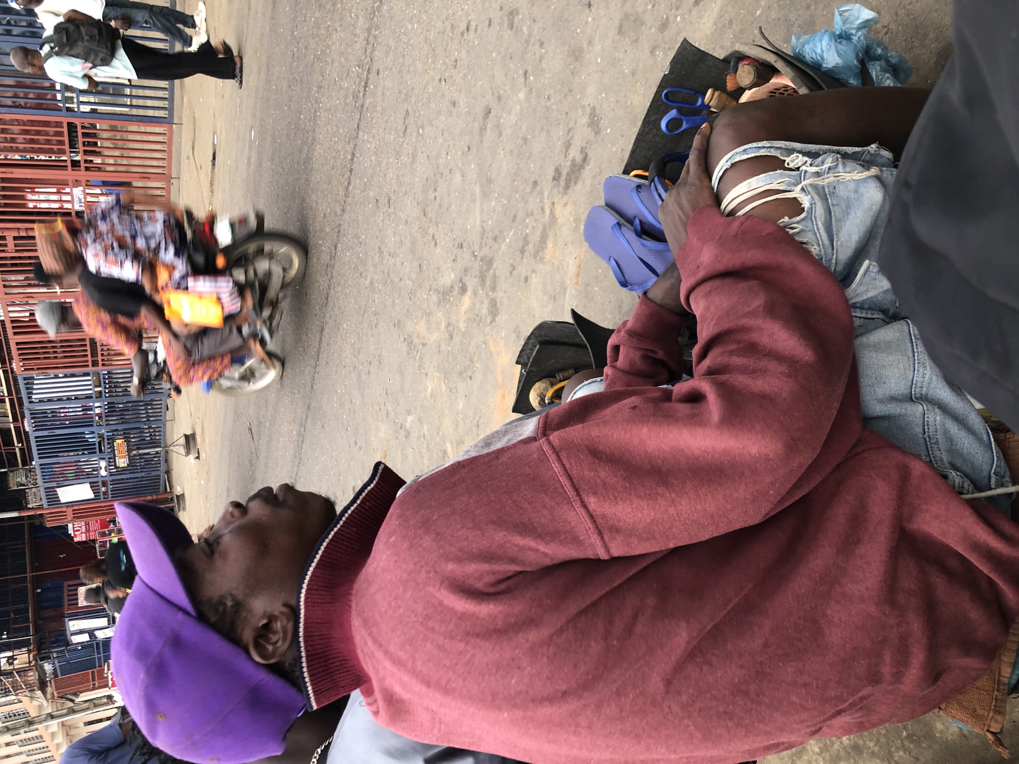 The image captures a street scene in Ikeja, Nigeria. In the foreground, a person wearing a purple baseball cap and a maroon sweatshirt is seated in what appears to be a small, portable station. The individual is wearing ripped denim shorts. To their right, a dark container holds various items, including a pair of blue flip-flops, a pair of blue scissors, and what looks like brushes. In the background, a motorcycle with three people on it is in motion, passing by a row of blue and red metal gates. Other individuals are visible in the distance, some walking and others also on motorcycles. The ground is paved, and the overall environment suggests a busy urban street.