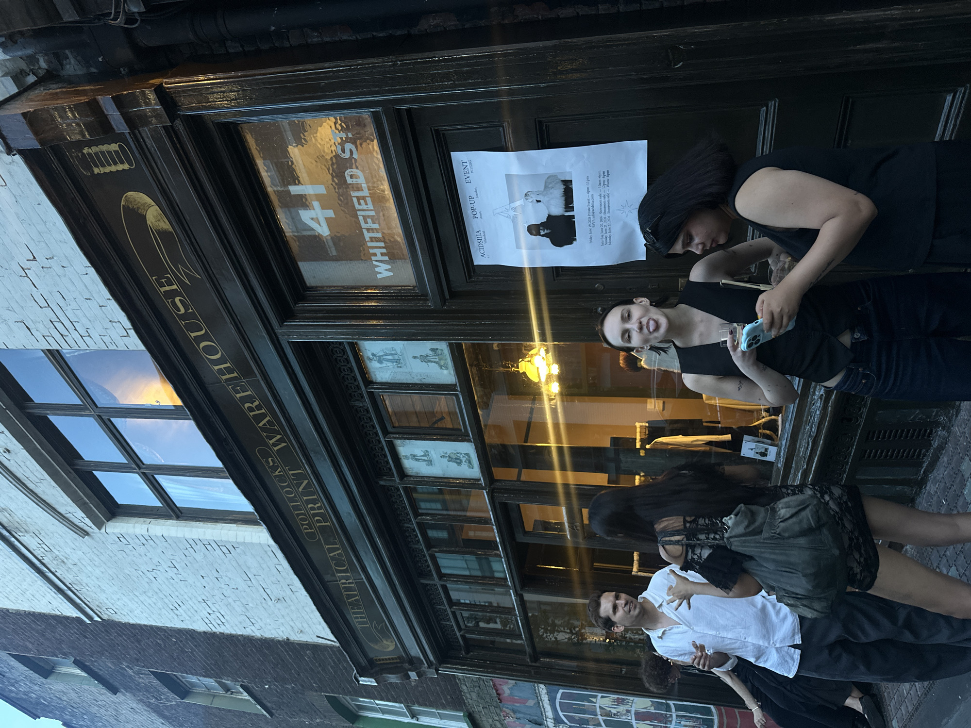 The image depicts a street scene in London, United Kingdom, outside Pollocks Theatrical Print Warehouse, located at 41 Whitfield Street. The building facade is dark wood with large glass windows. Above the windows, a sign reads "POLLOCKS THEATRICAL PRINT WAREHOUSE" in gold lettering. A poster for an "ACIDIIIIA POP-UP EVENT" is attached to the door.
Four individuals are present on the pavement in front of the building. On the left, a man in a white shirt is holding a glass. Next to him, a woman in a black lace dress with a large grey bag is facing away from the camera. In the center, a woman wearing a black vest and jeans has her tongue sticking out and is holding a drink. To her right, another woman in a black sleeveless top is looking at her phone. The lighting suggests it is late afternoon or early evening. The overall atmosphere appears casual and social, possibly at an event or gathering.