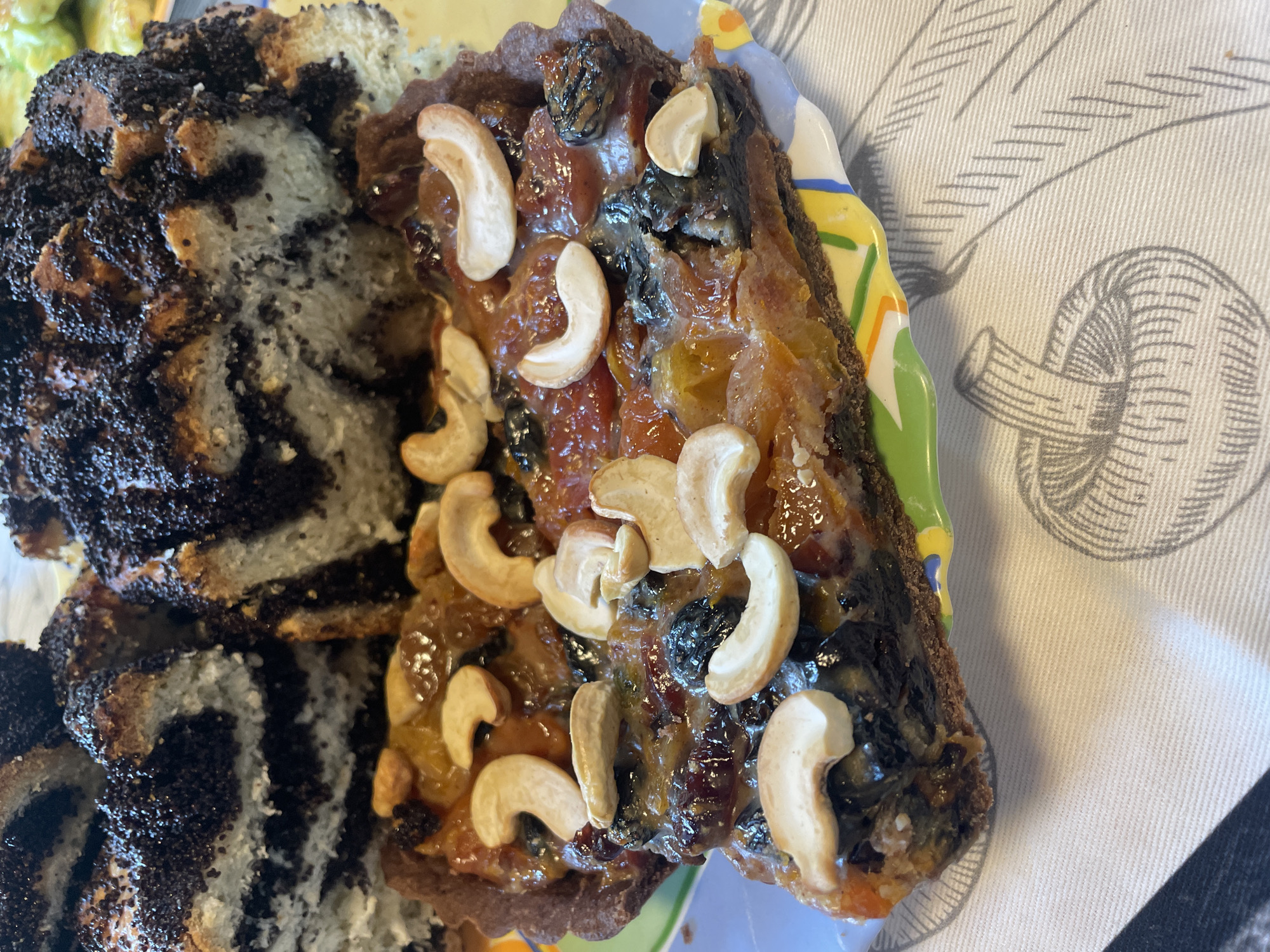 A close-up shot shows a slice of fruit tart with cashews, sitting in front of a poppy seed cake. The tart has a dark crust and is filled with an assortment of dried fruits, including raisins and possibly apricots, all coated in a glossy glaze. Several cashews are artfully placed on top. The poppy seed cake is visible in the background, with distinct layers of bread and poppy seed filling. The food is presented on a white tablecloth with a subtle pattern of apples. The setting appears to be indoors, likely a domestic kitchen or dining area. There is no indication of the specific location within the city of Турово, Russia.