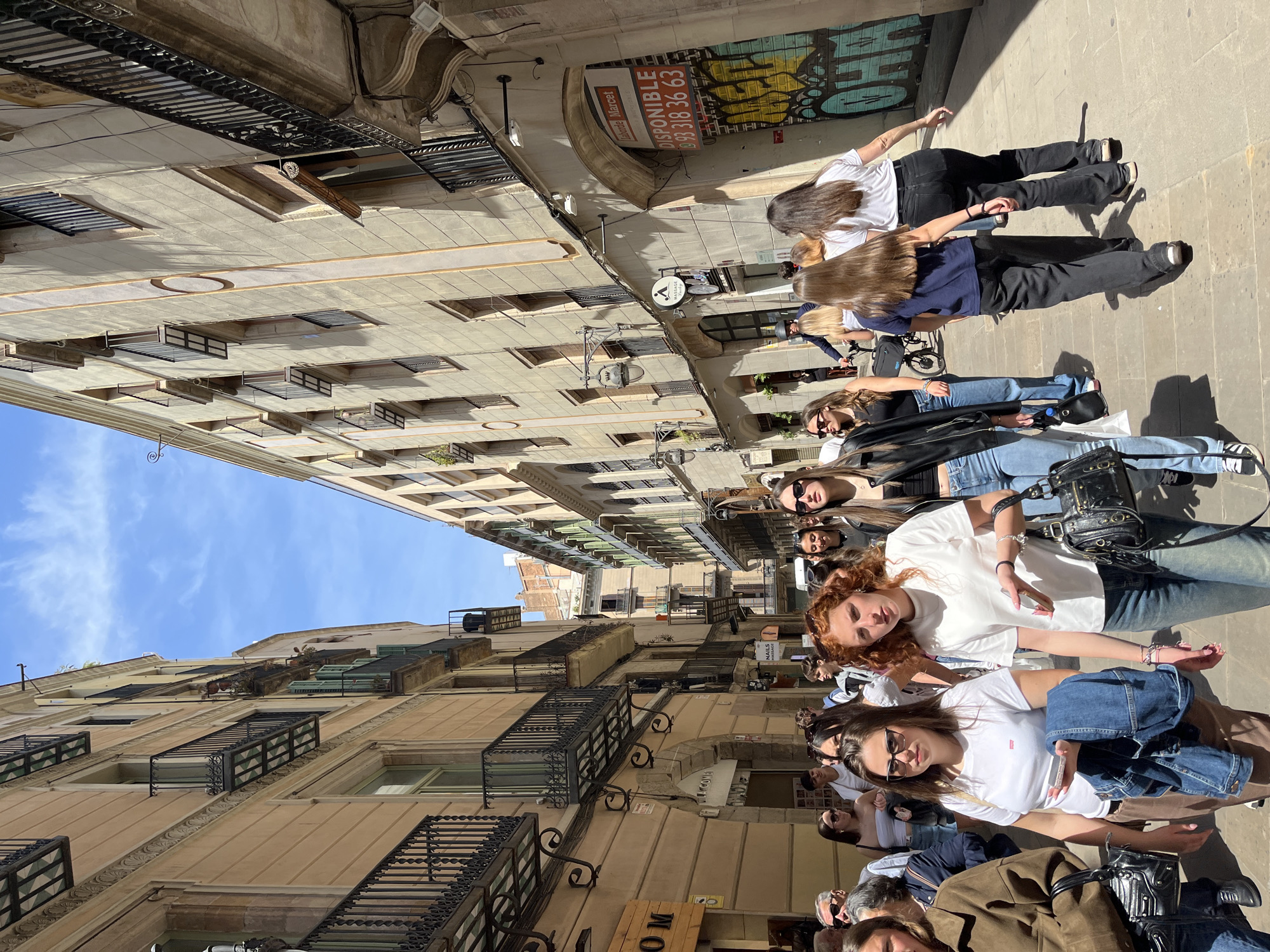 This vibrant street scene, set in Barcelona, Spain, captures a group of people walking and standing on a sunny day in a narrow urban alleyway. The image is taken from a slightly low angle, looking up towards the tall, traditional buildings that line both sides of the street. The architecture features multiple floors with ornate black wrought-iron balconies and various windows, some with roller blinds or shutters. A clear blue sky with a few wispy clouds is visible above the buildings, indicating bright daylight. Strong shadows are cast on the paved ground, suggesting a late morning or early afternoon setting.
The main subjects are a group of young people, predominantly women, dressed in casual modern attire including jeans, t-shirts, and sneakers. Many are wearing sunglasses. Some individuals in the foreground are looking directly at the camera with relaxed expressions, while others are seen from behind or profile as they move along the street. One pair of women further back appear to be holding hands. Among the objects visible are a black shoulder bag, a smartphone, and bicycles in the background.
Visible text includes a prominent red and white sign on a building with "DISPONIBLE" (Available) and a phone number: "93 318 36 63". Below this, colorful graffiti is present on a wall. Another sign reads "taberna Marcel," and a circular logo with the letter 'A' is visible on a storefront. A partial wooden sign in the bottom foreground shows "OM." The overall atmosphere is casual and lively, typical of a bustling European city street.