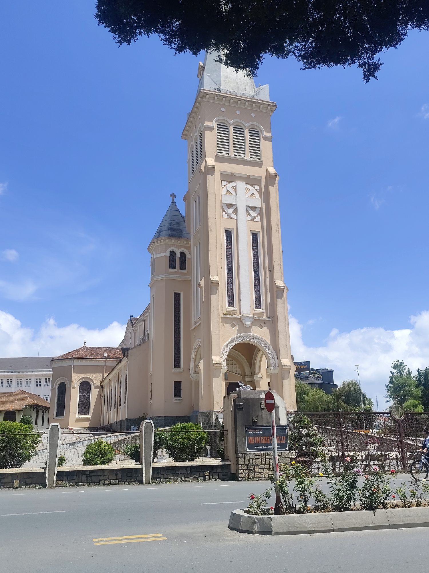 The image shows the Nha Tho Chinhtoa Da Lat cathedral, a Roman Catholic cathedral in Da Lat, Vietnam. The cathedral is a tall, beige-colored building with a prominent bell tower featuring a large white cross. It has arched windows and a conical roof on a smaller tower to the left. The cathedral is situated on a street with a road in the foreground, bordered by a low stone wall and greenery. A wrought-iron gate is visible to the right, with a sign in front of it. Several people are present, including one person riding a bicycle past the gate. The sky is blue with some scattered clouds.