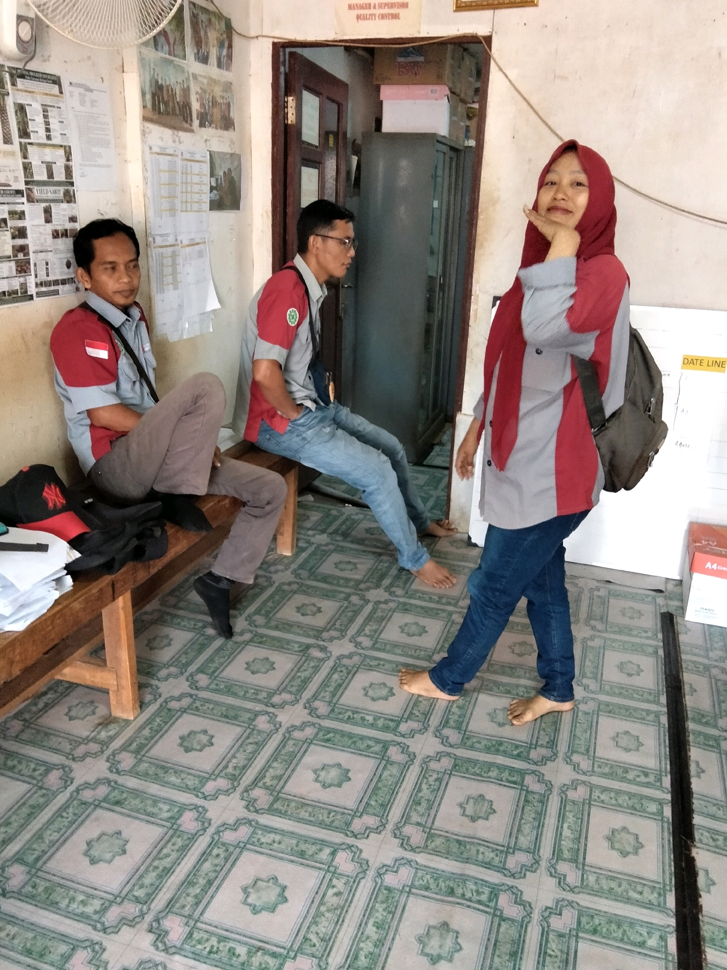 The image depicts an indoor scene, likely an office or waiting area, located in an unspecified region of Indonesia. Two men are seated on a wooden bench against a textured wall adorned with various documents and framed photographs. Both men are wearing collared shirts with red and grey accents, and one has a lanyard around his neck. The man on the left is looking to the side, while the man on the right is looking forward. A stack of papers and a black baseball cap with a red logo are placed on the bench.
In the foreground, a young woman wearing a red hijab and a grey and red collared shirt stands facing the viewer. She is wearing blue jeans and is barefoot. Her left hand is raised to her chin, and she appears to be posing for the photo. A dark-colored backpack is slung over her right shoulder.
The room's floor is covered in a patterned tile design featuring green and pink geometric shapes. A metal filing cabinet and a door are visible in the background. A whiteboard with the text "DATE LINE" is mounted on the wall behind the woman. The overall impression is of a casual, everyday setting within a workplace.