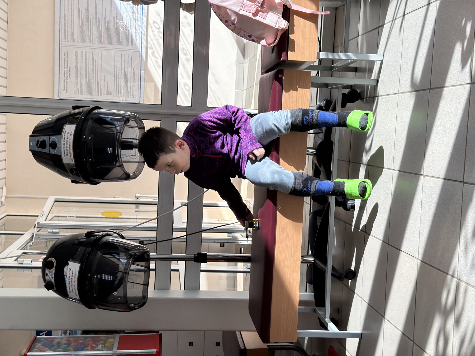 A young boy, dressed in a purple long-sleeved shirt, light grey pants, and dark socks with bright green foot attachments, lies on his back on a padded wooden bench. He appears to be engaged in physical therapy or exercise, with his feet connected to some form of rehabilitation equipment. His left hand is reaching towards a strap or part of the apparatus.
The setting is an indoor space, likely a facility such as a rehabilitation center or a specialized gym in Chaikovsky, Russia. Bright sunlight streams in through large windows on the left, casting distinct shadows on the light-colored tiled floor and walls.
To the boy's left, two black, helmet-like devices on stands are prominent. These devices have control knobs and a clear visor. Visible text on a sticker on one of these devices reads in Russian: "Уважаемые клиенты! После сушки волос выключайте сушуары!!!" which translates to "Dear clients! After drying your hair, turn off the dryers!!!" This indicates they are hair dryers.
In the background, a pink backpack rests on another bench or surface. Further text is faintly visible on a poster in the background, including "Правила регулирования расписания..." (Rules for regulating the schedule...) and "учреждения" (institutions/establishments). The word "COWAY" is also visible on one of the boy's socks. The overall scene suggests a functional, well-lit environment.