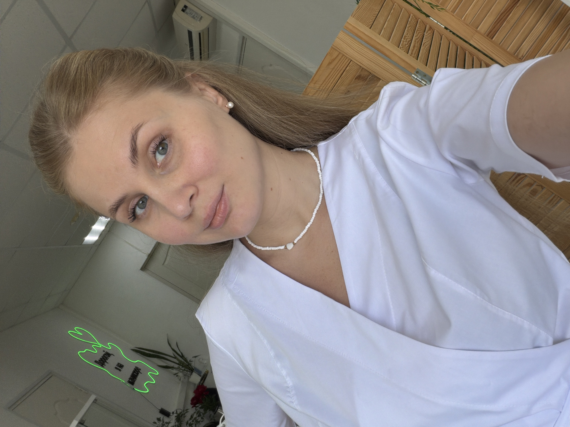 The image displays a selfie of a young woman, likely taken as a reflection in a mirror or glossy surface, indicated by the inverted background elements and the camera angle. The woman has fair skin, light blonde hair pulled back, and light blue-grey eyes, gazing directly at the viewer with a slight, calm smile. She wears a white, professional-looking V-neck uniform top, possibly a medical scrub or spa attire. Her accessories include small pearl stud earrings and a white beaded necklace with a central heart-shaped charm.
The background, seen through the reflection, reveals an indoor setting that appears to be a commercial space. A prominent feature is a bright green neon sign outlining Cyrillic text, which reads "Дорогой маникюр" (Dear manicure or Expensive manicure). This text strongly suggests the location is a beauty salon, nail studio, or similar establishment. Other visible elements in the reflection include a white ceiling, an air conditioning unit, parts of white walls and doorways, various potted plants (one with red flowers, another with green leaves), and a wooden slatted screen or divider. The environment appears clean, well-lit, and professionally maintained, consistent with a service-oriented business in Chaikovsky, Russia.