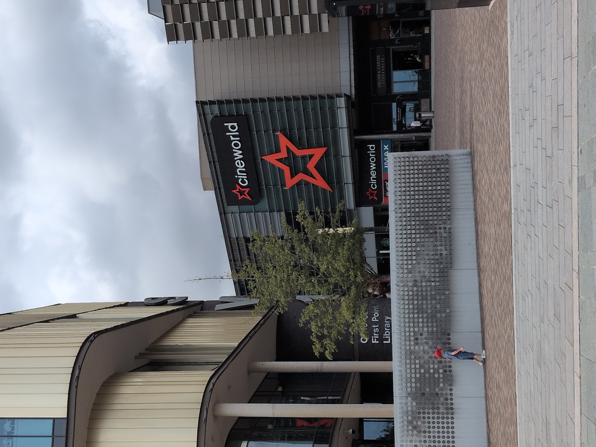 A boy wearing a red cap and blue shorts walks along a paved walkway in front of a modern building complex. To the left, a building with cream-colored vertical siding curves around a corner, with large windows visible. Further to the right, a larger building features a large "cineworld" sign with a red star logo. Below this, another "cineworld" sign is visible along with "IMAX." Adjacent to the cineworld entrance is a Miller & Carter Steakhouse. A tree stands between the cineworld and the library. The ground in front of the buildings is paved with bricks, and a decorative metal wall with a circular pattern runs along the walkway. The sky is overcast. The scene appears to be the Southwater area of Telford, United Kingdom, a leisure and entertainment district.