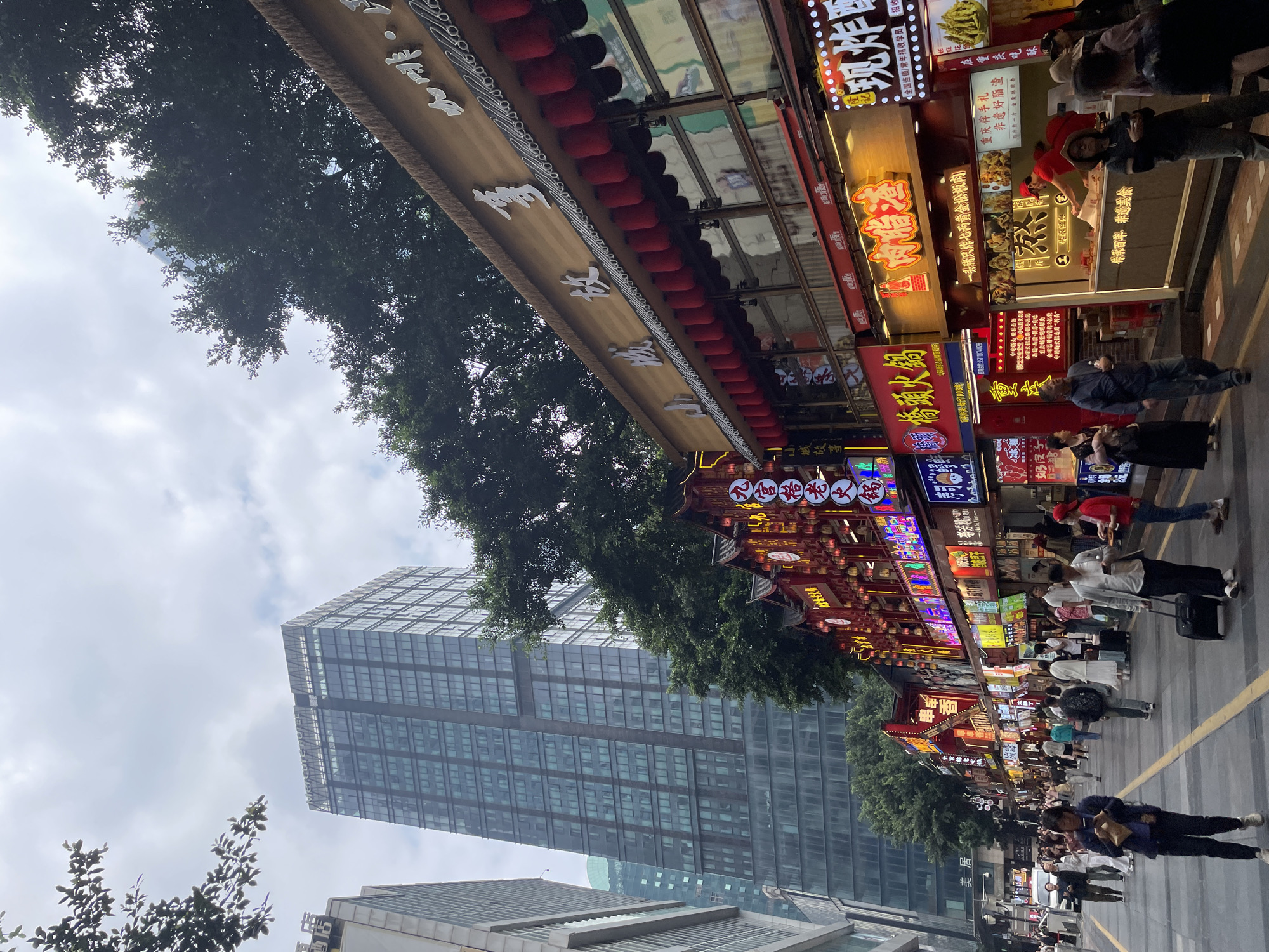 The image depicts a bustling street scene in Yuzhong District, China. A tall, modern skyscraper rises in the background, contrasting with the traditional Chinese architecture lining the street. Numerous shops and restaurants are illuminated with bright neon signs in Chinese characters, adding to the vibrant atmosphere. Many pedestrians populate the street, engaging in various activities such as walking, shopping, and dining. Red lanterns and other decorative elements enhance the traditional feel of the area. The scene suggests a blend of modern urban life with traditional Chinese culture.