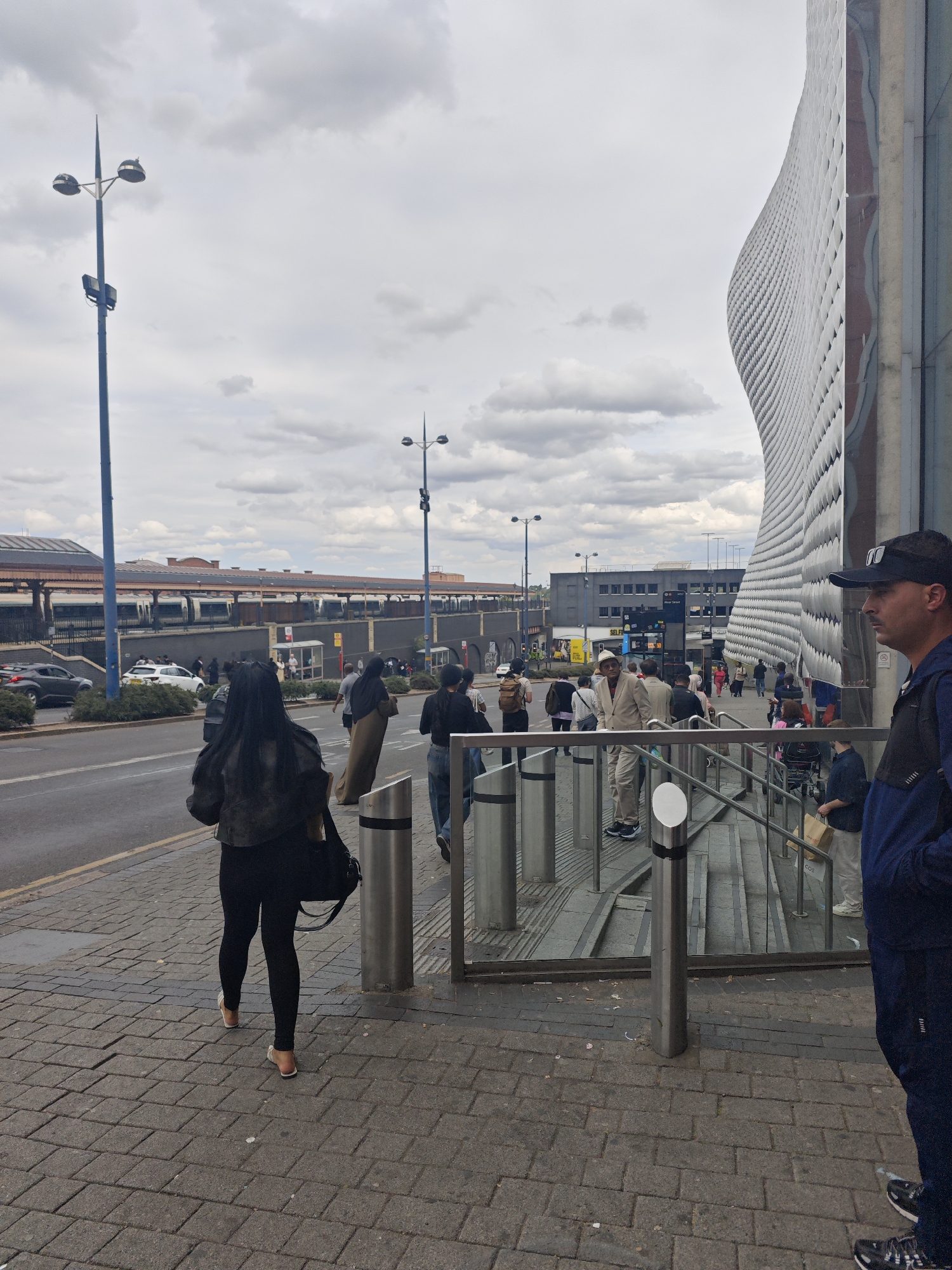 This is a ground-level, eye-level, full shot taken outdoors on a cloudy day in Birmingham, United Kingdom, in front of the Bullring shopping centre. The facade of the Bullring shopping centre, characterized by its curved, patterned white exterior, dominates the right side of the frame. In the foreground, a man wearing a navy blue jacket and a black baseball cap stands facing towards the right. To the left of the man, a set of stairs with metal railings leads down. Several people are visible walking on the pavement and towards the stairs. Further to the left, a street with traffic, including a white car and a black car, runs parallel to the pavement. In the background, a train station with a long platform and trains is visible, along with a blue lamppost. The ground is paved with grey cobblestones.