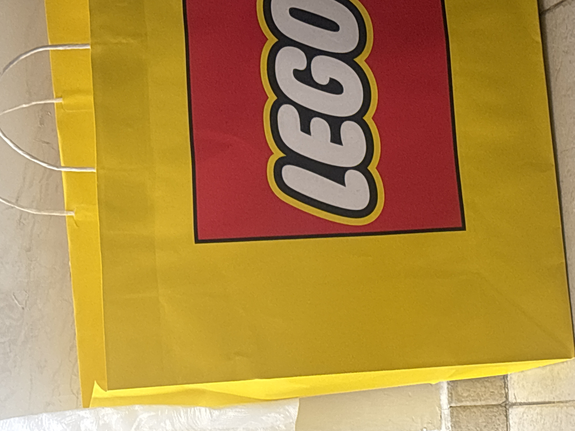 A close-up photograph depicts the upper portion of a yellow LEGO paper shopping bag with a red rectangular LEGO logo in black and white. The bag has white string handles. The photograph was taken indoors, on a light tile surface. There is no indication of the location being San Pablo del Monte, Mexico. No people, subjects, actions or interactions are visible in the image.