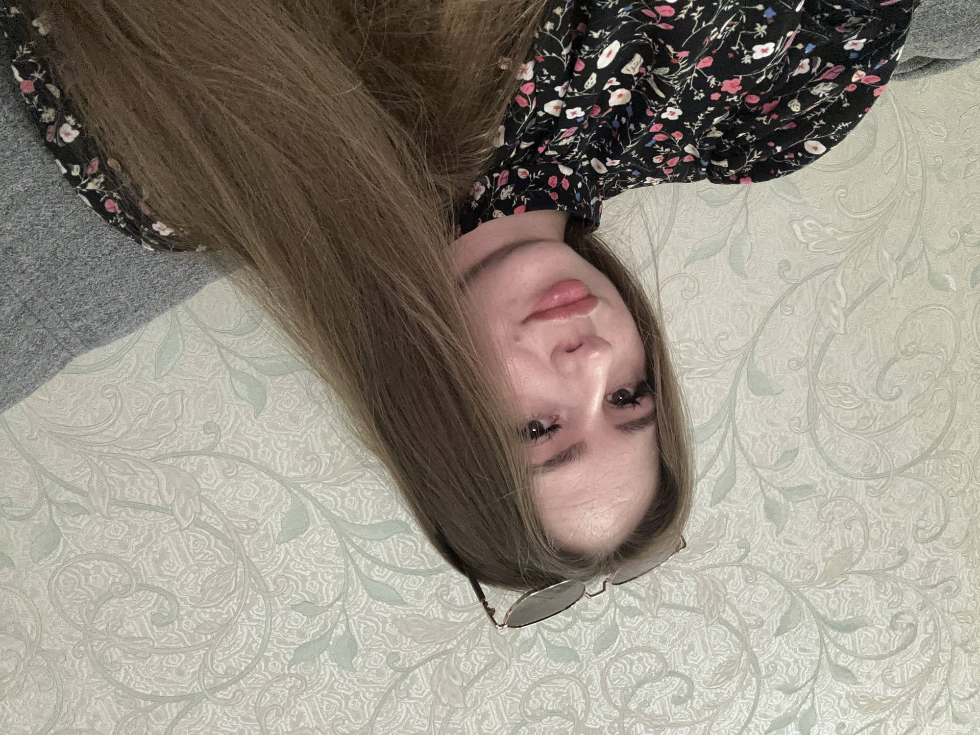 A young woman with long, brown hair and sunglasses perched on her head is pictured from the chest up. She is wearing a black dress with a floral pattern of small pink and white flowers. Her eyes are looking forward, and her lips are slightly parted. The background is a light-colored wallpaper with a subtle floral pattern. She is sitting on a grey couch or chair. The lighting appears to be indoor.