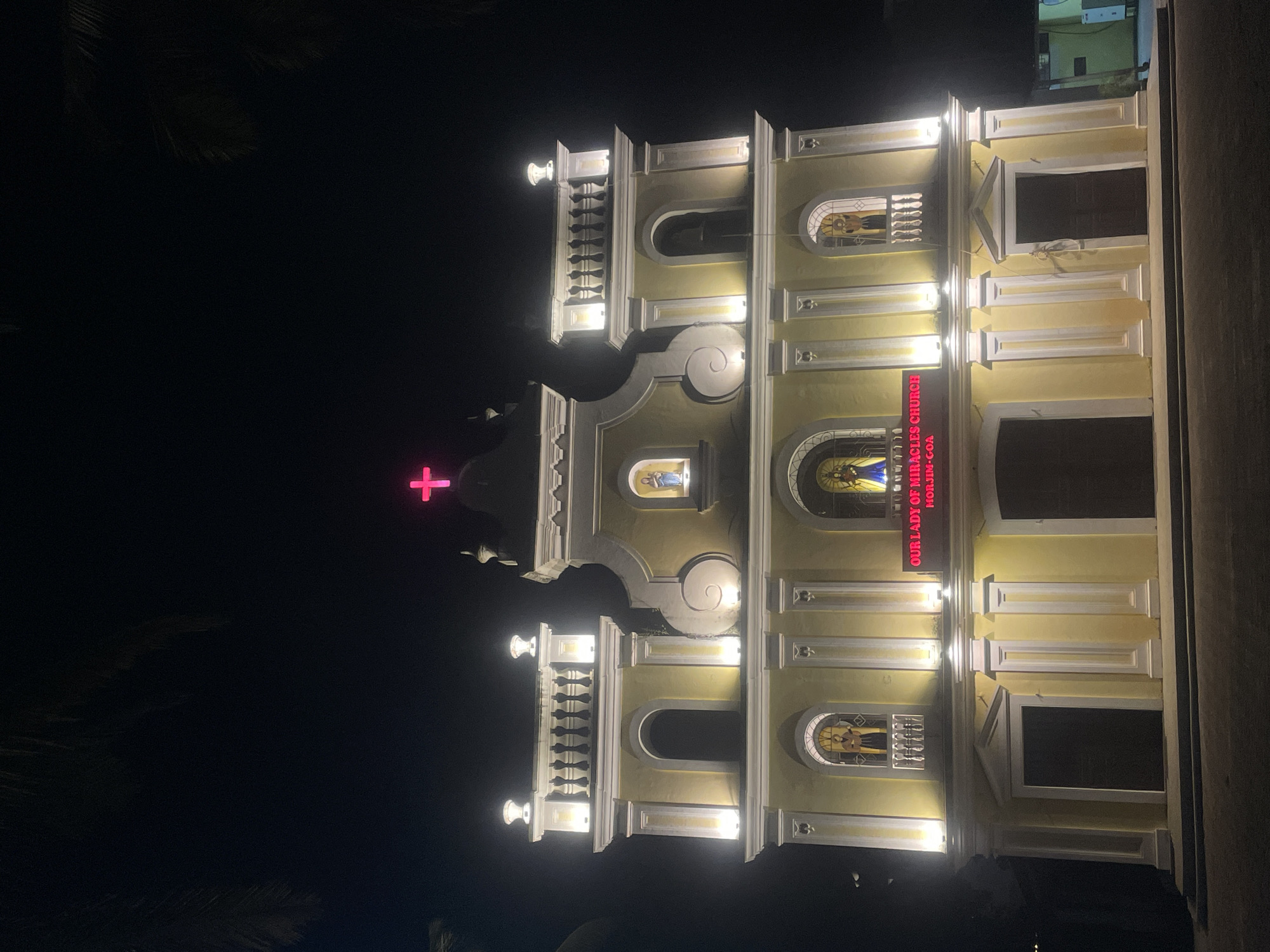 The image displays the illuminated facade of "Our Lady of Miracles Church" at night. The church building, painted in a light yellow or cream color, stands out brightly against the dark sky. Its architecture features classical elements, including decorative moldings, columns, and cornices, with multiple levels and sections.
Strategically placed white artificial lights highlight the church's architectural details, casting a warm glow on the structure. Two prominent balconies with white balustrades are visible on the upper sections of the central facade.
At the highest point, a vibrant neon pink cross glows prominently. Below it, within an arched niche, a small, illuminated statue or religious figure can be seen. Further down, several arched windows contain what appear to be stained-glass depictions or statues. One such window on the lower right (as rotated) shows a figure holding what looks like a guitar. Another arched window on the upper left features a blue-robed figure.
On the right side of the main facade, a large red sign with white lettering clearly reads: "OUR LADY OF MIRACLES CHURCH MORJIM-GOA". The overall impression is one of peaceful nocturnal illumination, emphasizing the church's grandeur and sacred presence. No people or activities are visible, presenting a static nighttime scene. The location is Morjim, Goa, India, within the broader context of Ashvem.