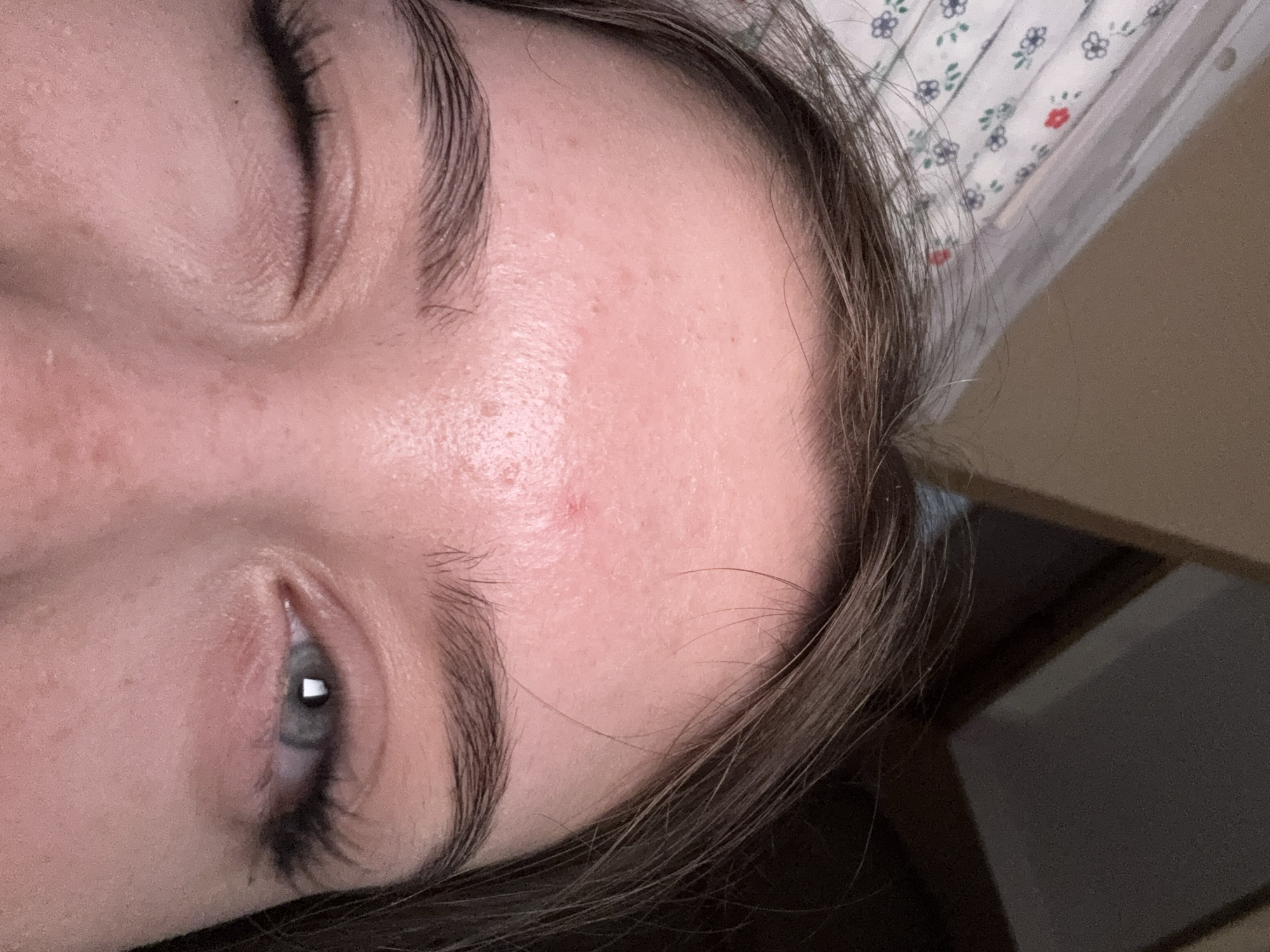 A close-up, eye-level shot focuses on the forehead and eyes of a young person. The subject is looking at the camera with their left eye open and their right eye winking. The left eye is a striking blue-gray, reflecting a light source. The right eye is partially closed. Both eyes are adorned with dramatic, long false eyelashes. The subject's eyebrows are dark and well-defined. The skin on the forehead appears fair with a scattering of freckles and some minor redness. A small red blemish is visible on the forehead, slightly to the left of center. The background is softly blurred, showing what appears to be a patterned fabric, possibly a sheet, on the left and a darker, indistinct area on the right. The lighting is dim, casting a soft glow on the subject's face. The image is taken from a head-on perspective, with the camera positioned slightly above the subject's eye level.